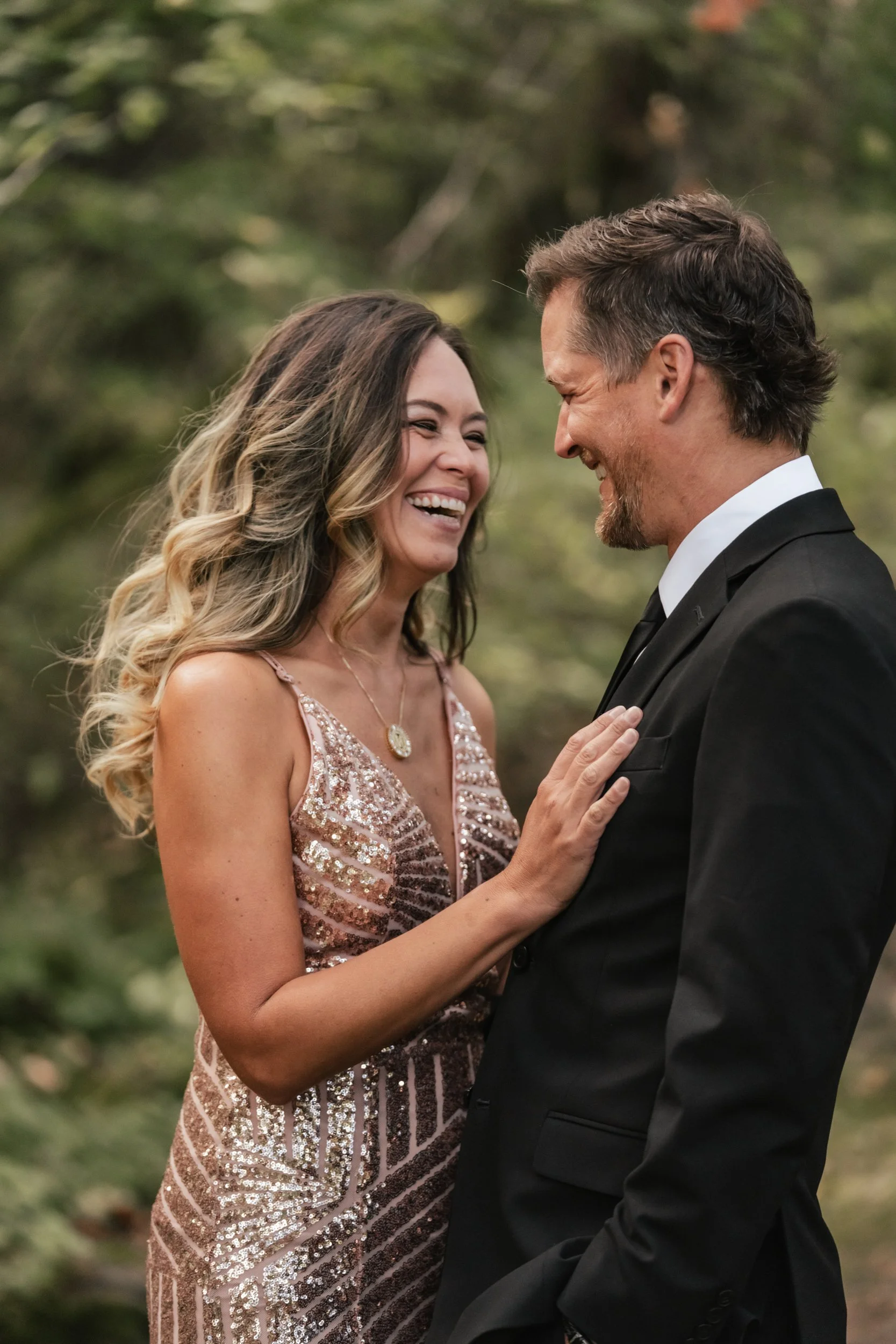 A woman in a fancy dress laughs authentically as she touches the chest of her husband wearing a suit.