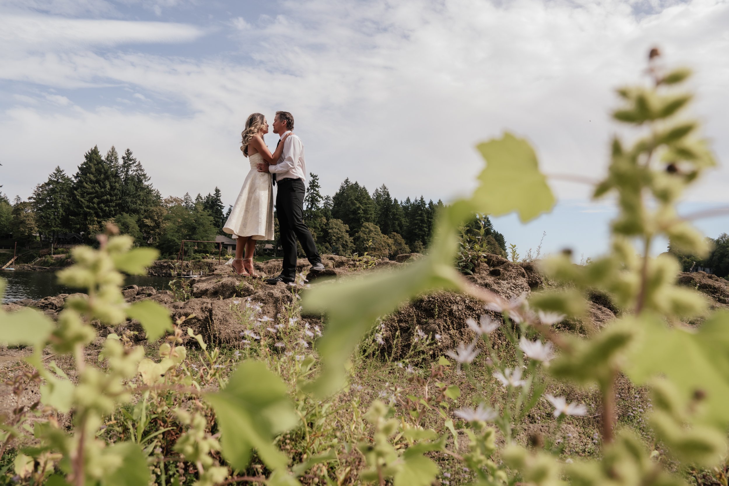 A couple hold each other tightly on top of a rock with foliage in the foreground and background.