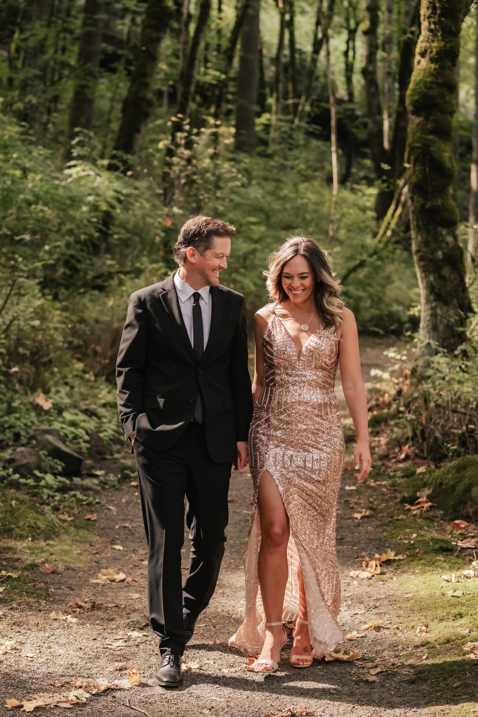 A man in a full suit and a woman in a gorgeous dress walk happily side by side at Tryon State Park.
