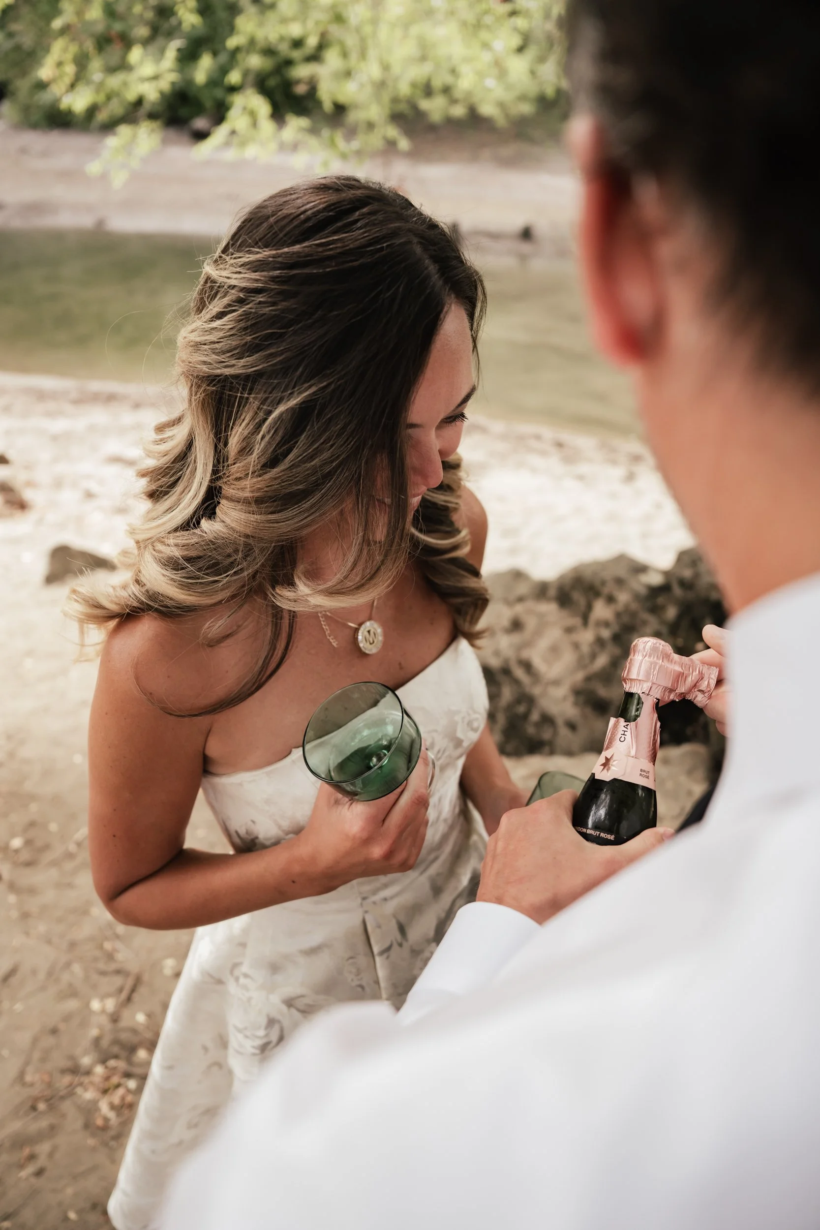 A husband prepares a celebratory drink for his wife on a sandy shoreline.