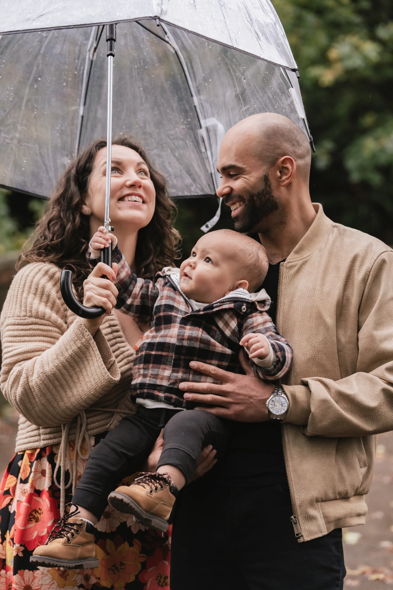 A mom and dad laugh at their baby trying to hold a clear umbrella.