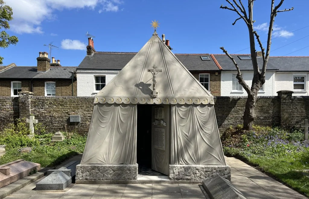 A small tent-shaped silver structure with gold decorative elements, a cross and crucifix on top and a window with a sign inside, set in a graveyard with graves and a brick wall in the background.