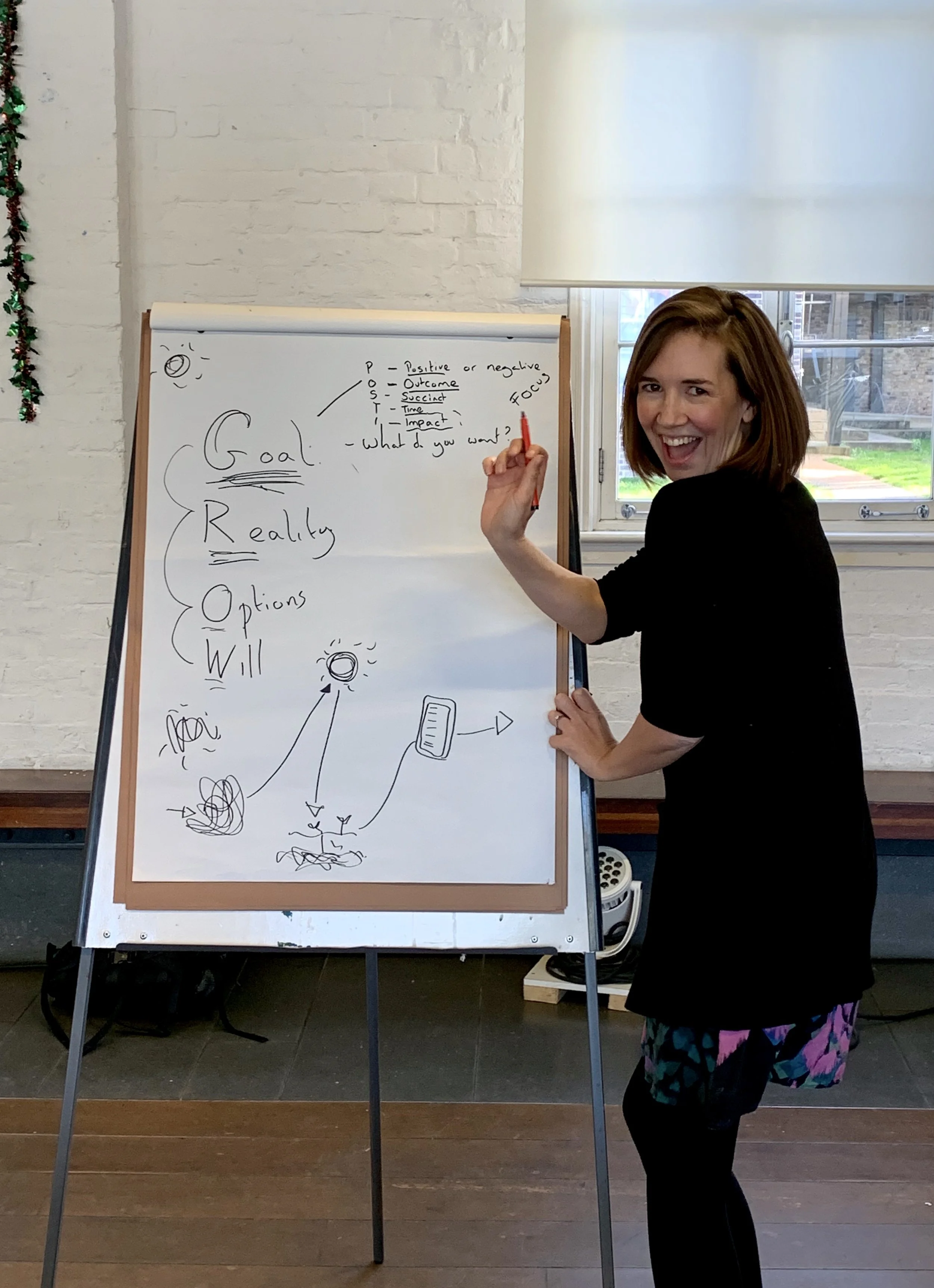 A woman with shoulder-length hair smiling and holding a red marker, standing beside a whiteboard with handwritten notes and drawings, in a room with a window and brick wall.