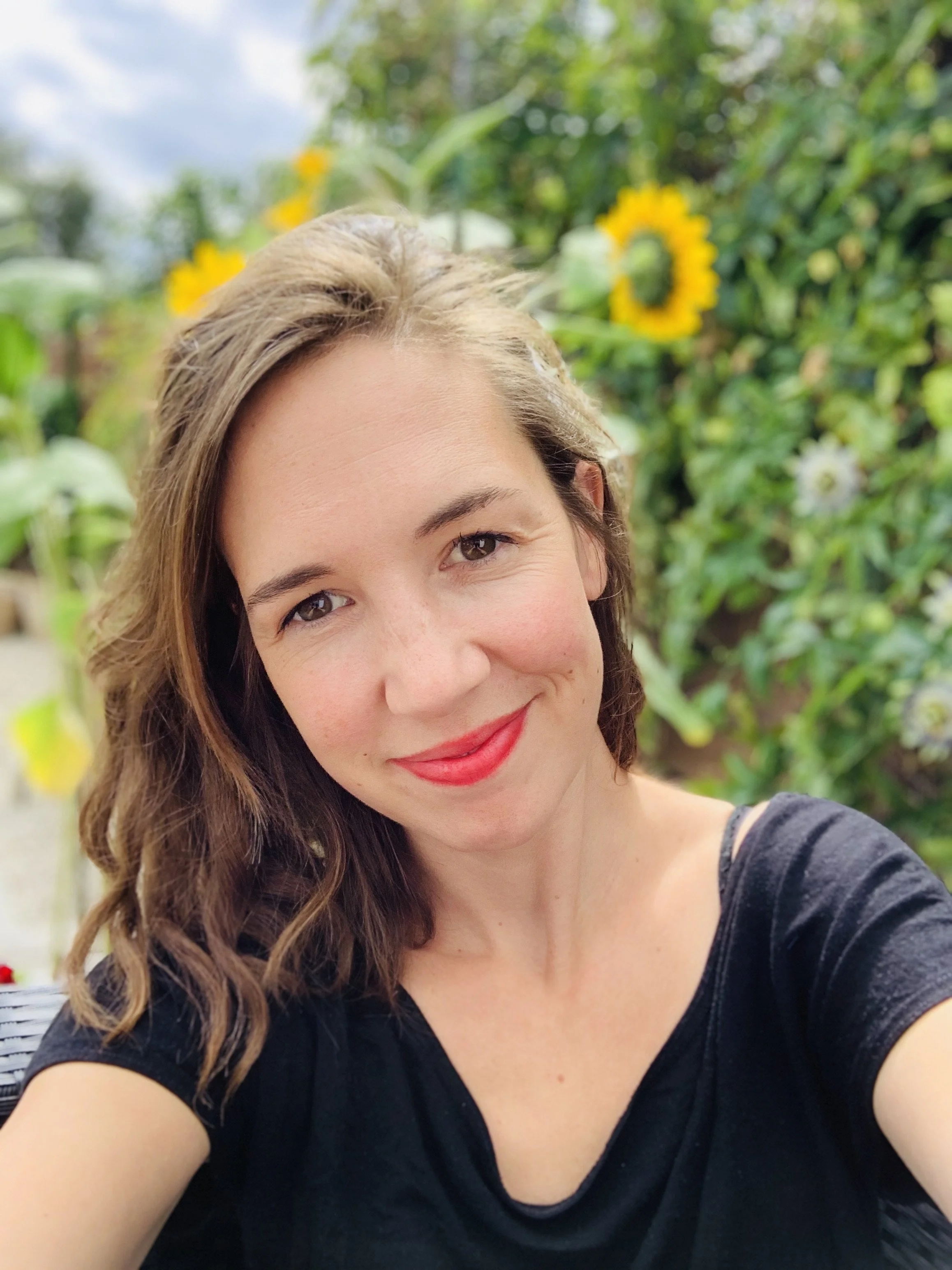 Close-up of a smiling woman with wavy brown hair and red lipstick in front of a garden with yellow sunflowers.
