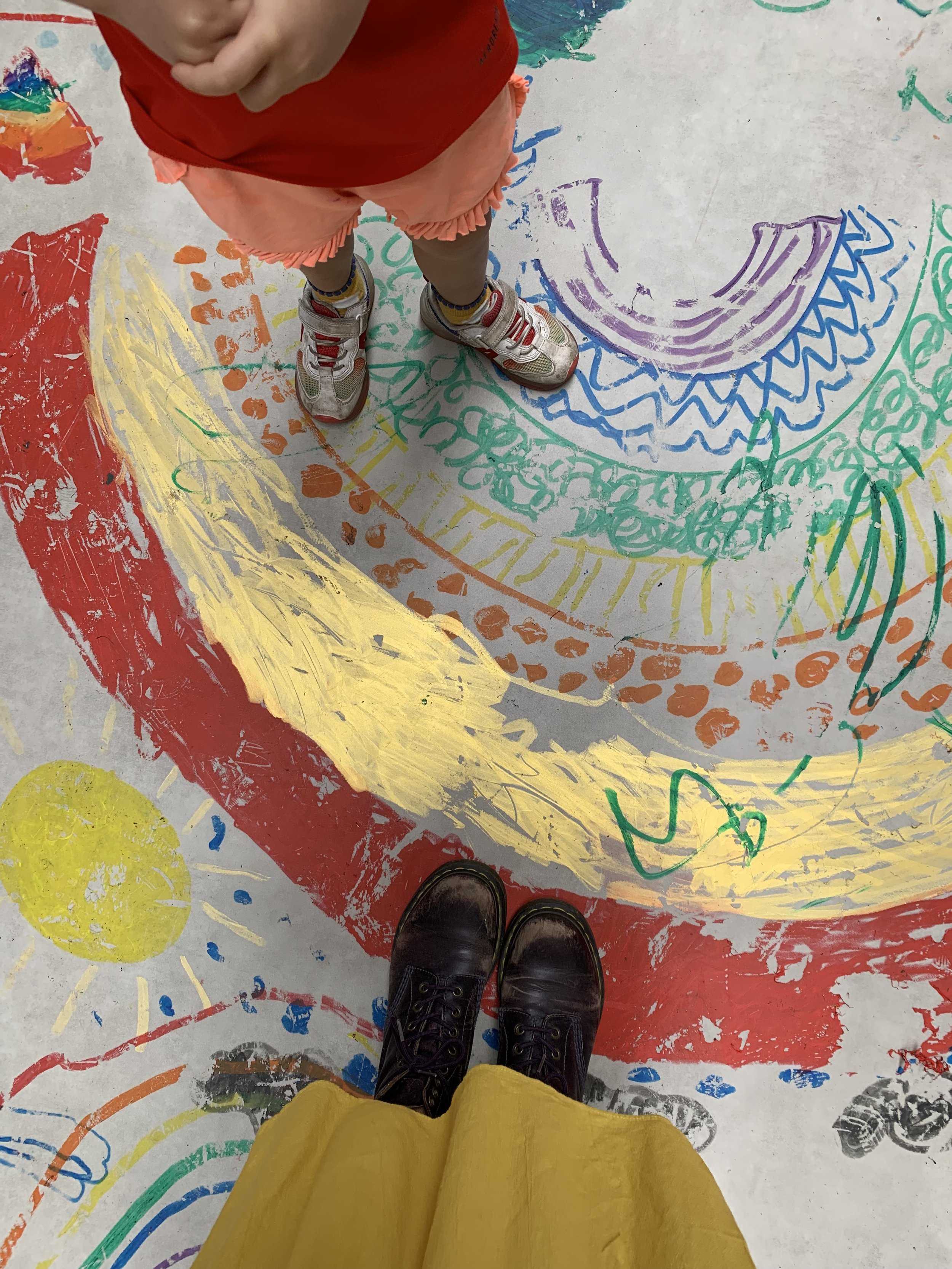View of a person's feet wearing black shoes standing on a colorful, painted floor with various abstract patterns and designs in red, yellow, green, blue, purple, and orange.