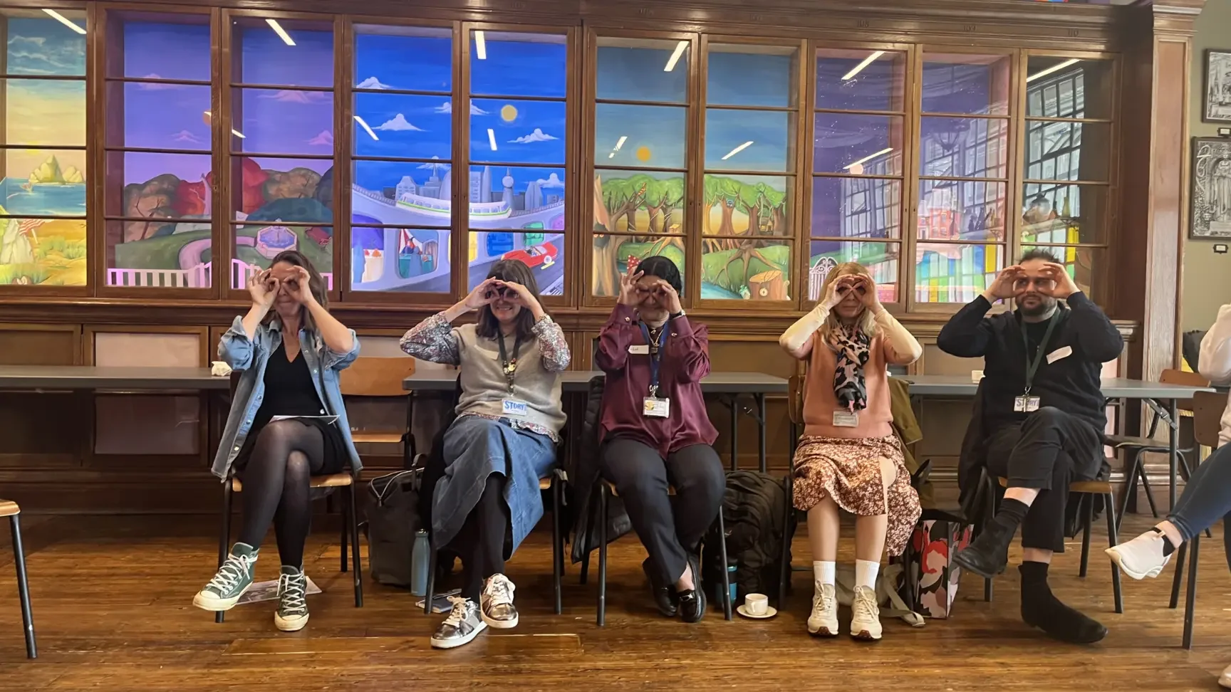 Five people sitting side by side on chairs indoors, each making a circular shape with their hands around their eyes, facing forward. Behind them is a colorful, painted mural depicting various scenes including a city skyline, trees, mountains, and a sunrise or sunset, within a wooden frame.