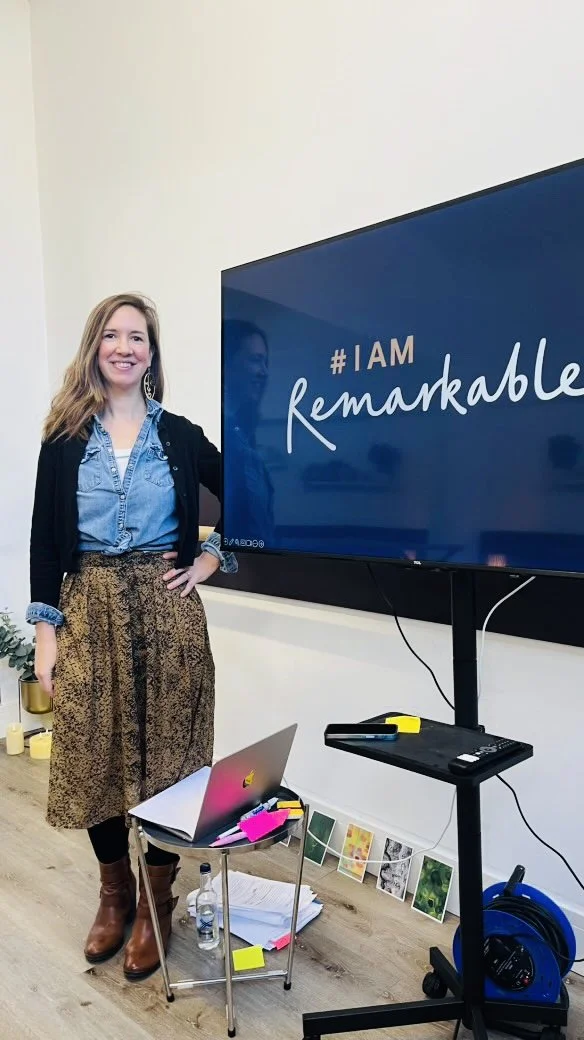 A woman standing next to a large screen that says '#I AM remarkable.' She has long hair, is smiling, and is dressed in a denim shirt, black cardigan, and a patterned skirt with brown boots. She is in a room with a table holding a laptop, water bottle, and some papers and markers. There are small potted plants and artwork on the floor against the wall.