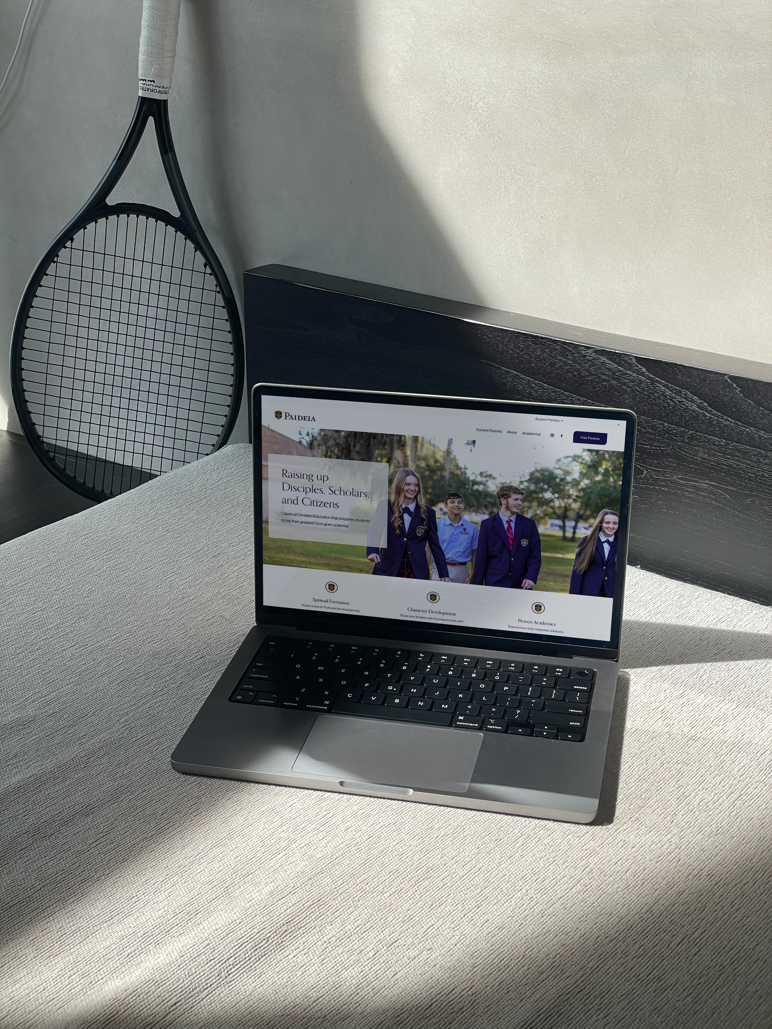 A laptop on a desk displaying a website for a school called Paideia, with four students in uniforms outdoors on the screen. A tennis racket leans against the wall in the background.