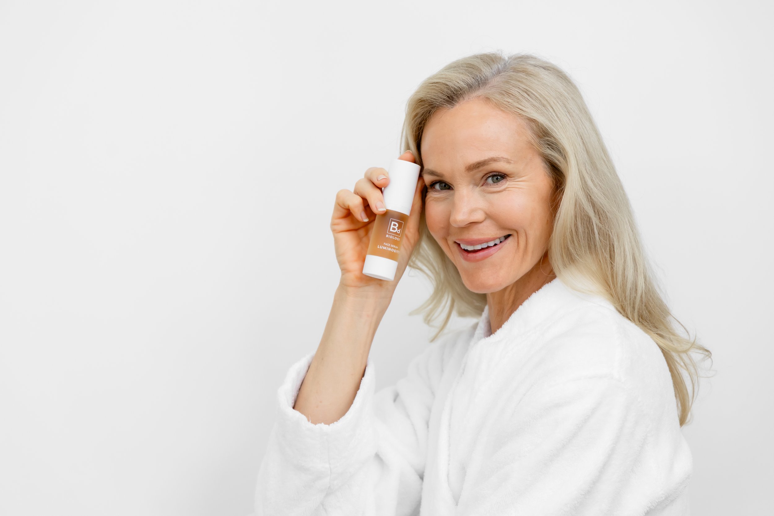Smiling woman with blonde hair in a white bathrobe holding a bottle of skincare product against a plain white background.