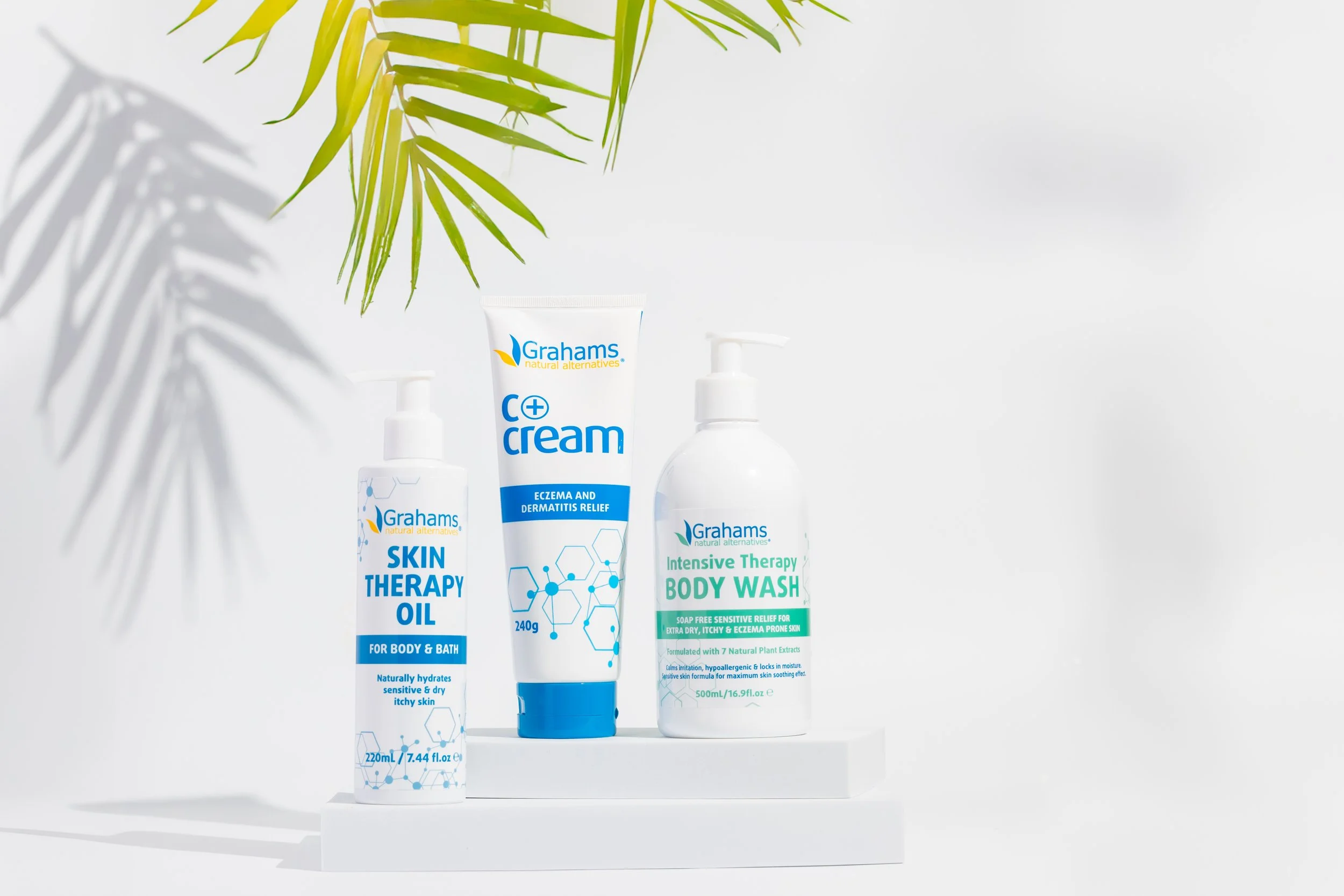 Three skincare products from Grahams arranged on a white surface with a white background; a skin therapy oil, an eczema and dermatitis relief cream, and an intensive therapy body wash, partially shaded by green palm leaves.