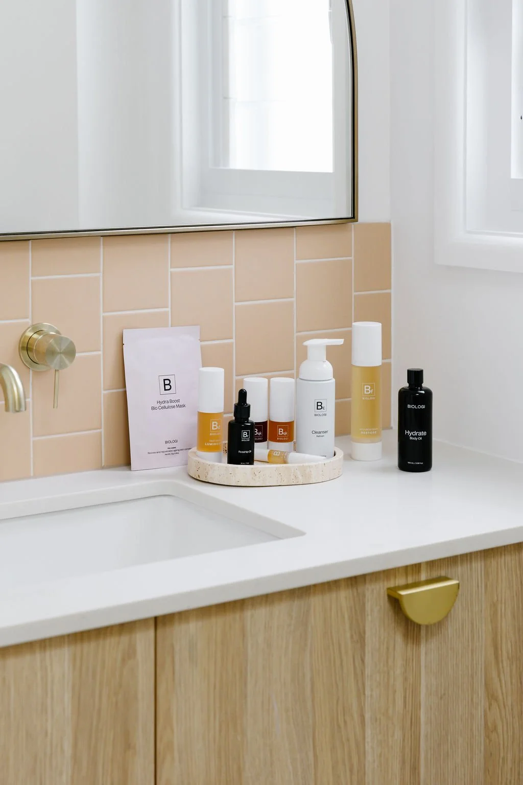 Bathroom vanity with assorted skincare and haircare products on a round tray, beige tiled wall, mirror, and window.