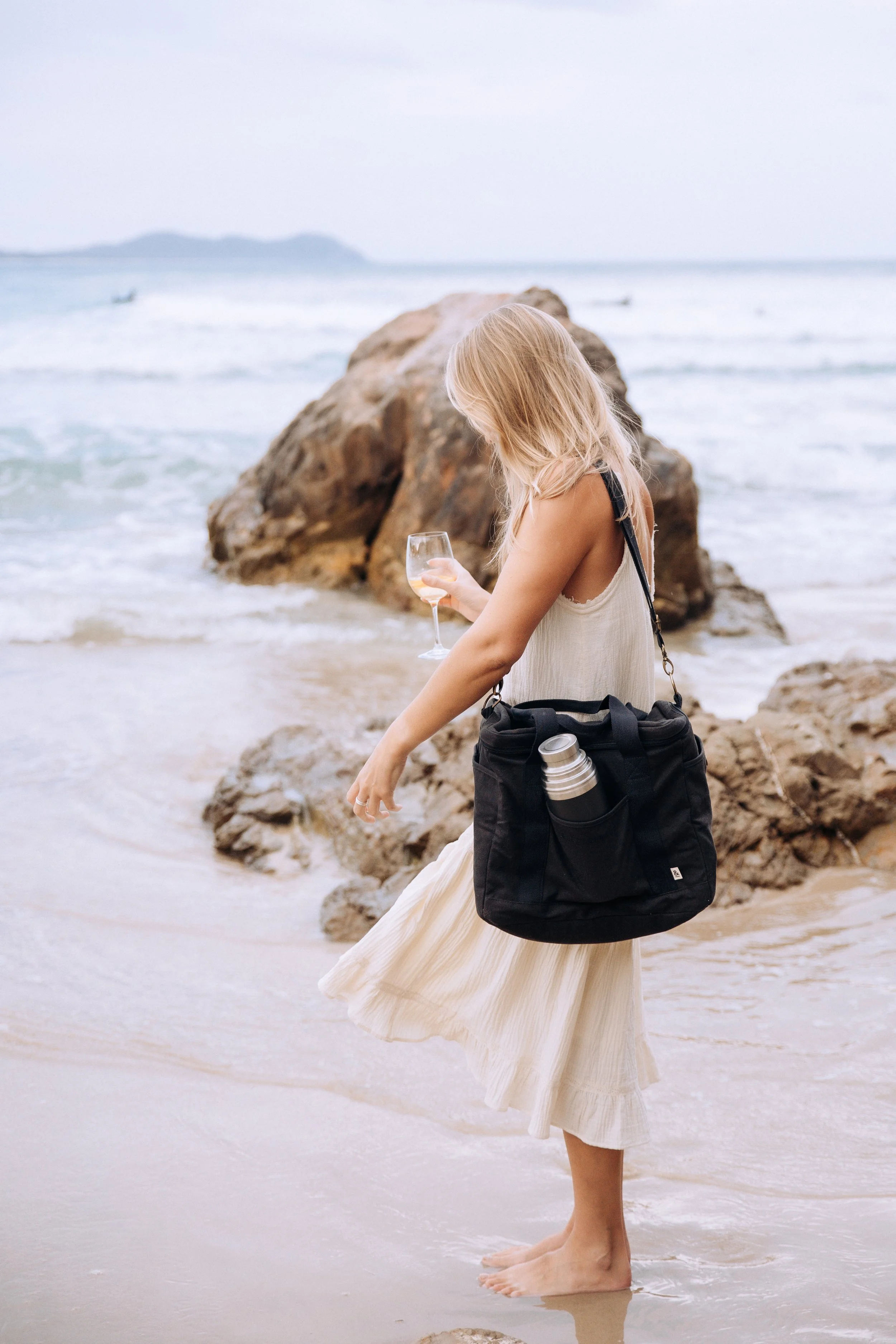A woman with blonde hair standing barefoot on the beach, holding a glass of white wine in one hand and a black bag with a water bottle in the other. She is wearing a light-colored dress, near rocks with the ocean in the background.