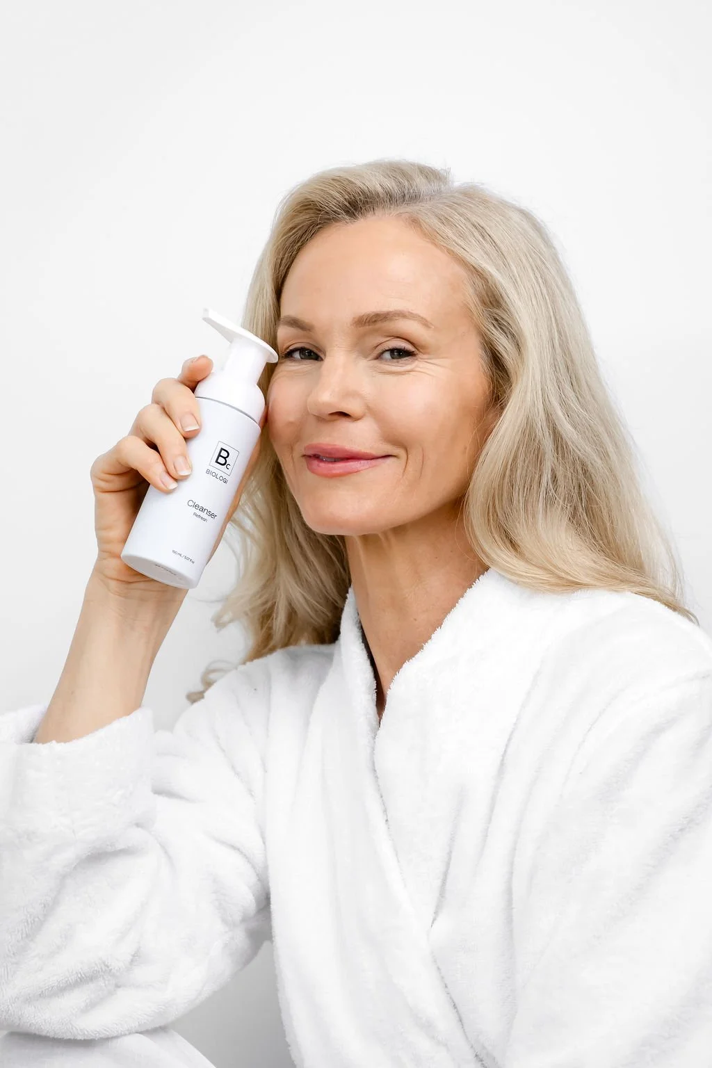 A smiling woman with blonde hair in a white bathrobe holding a facial cleansing device against her face.