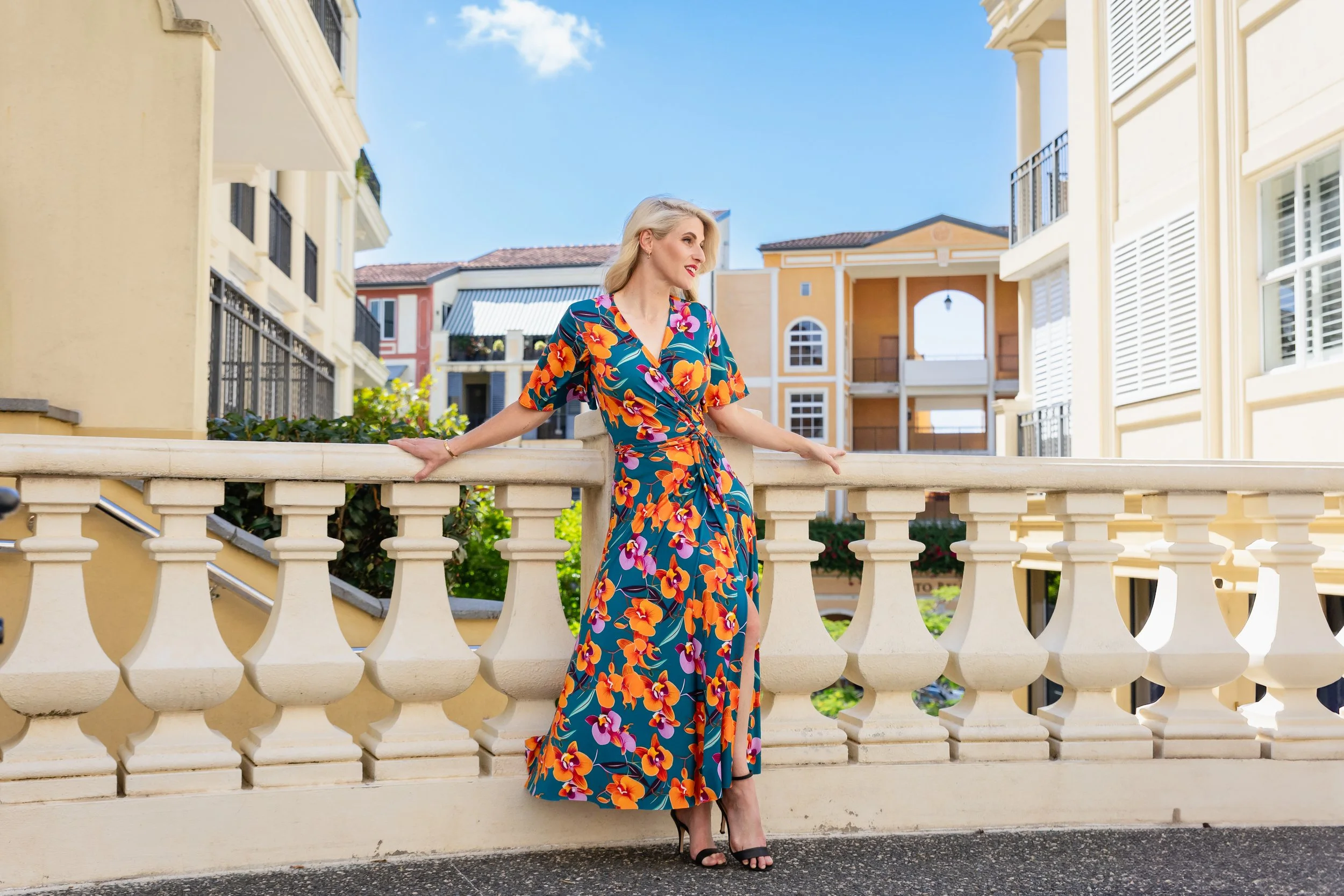 A woman in a colorful floral dress and black heels standing on a decorative white balcony with buildings in the background on a sunny day.