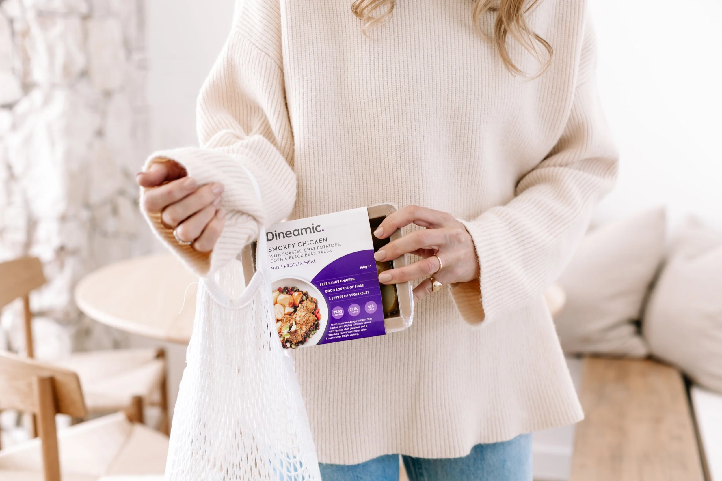 Person holding a plastic container with smoked chicken, a white net bag, and a gold ring on the finger in a bright room.