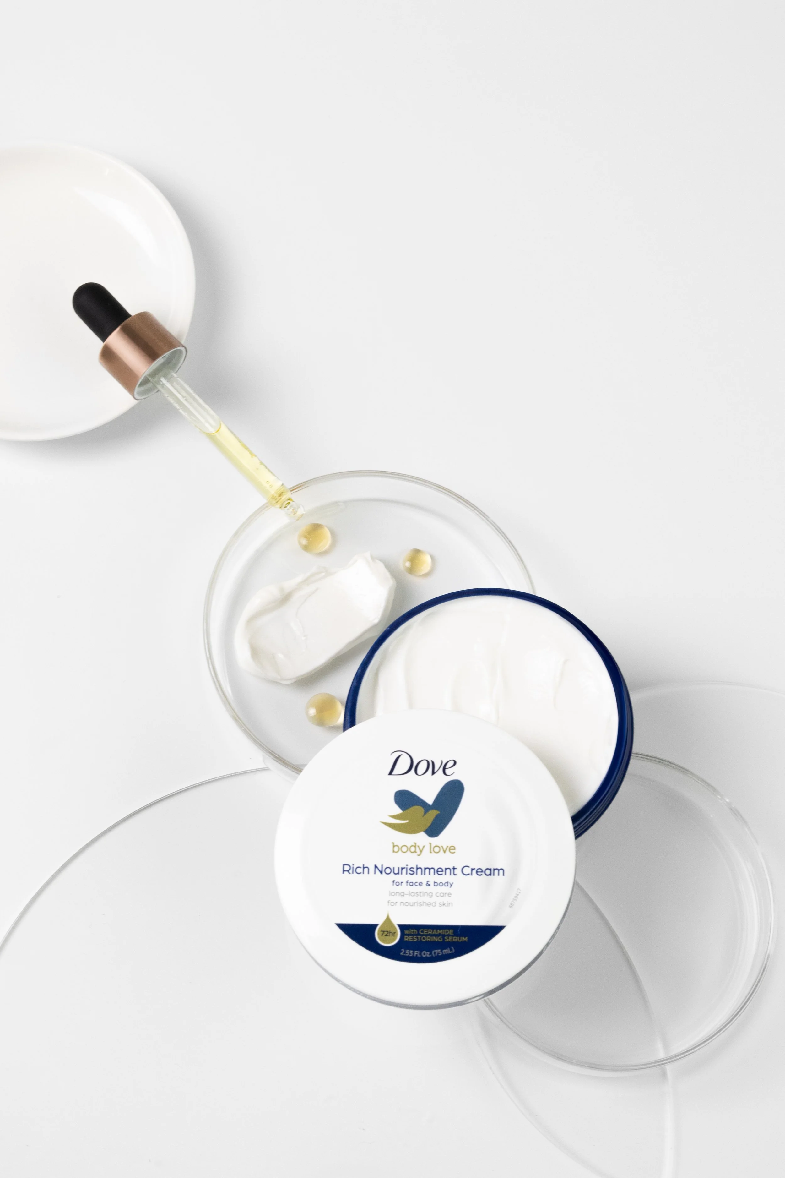 A jar of Dove Rich Nourishment Cream body and face moisturizer, an open container of cream, a glass dish with white cream, a dropper with liquid, and empty glass containers on a white surface.