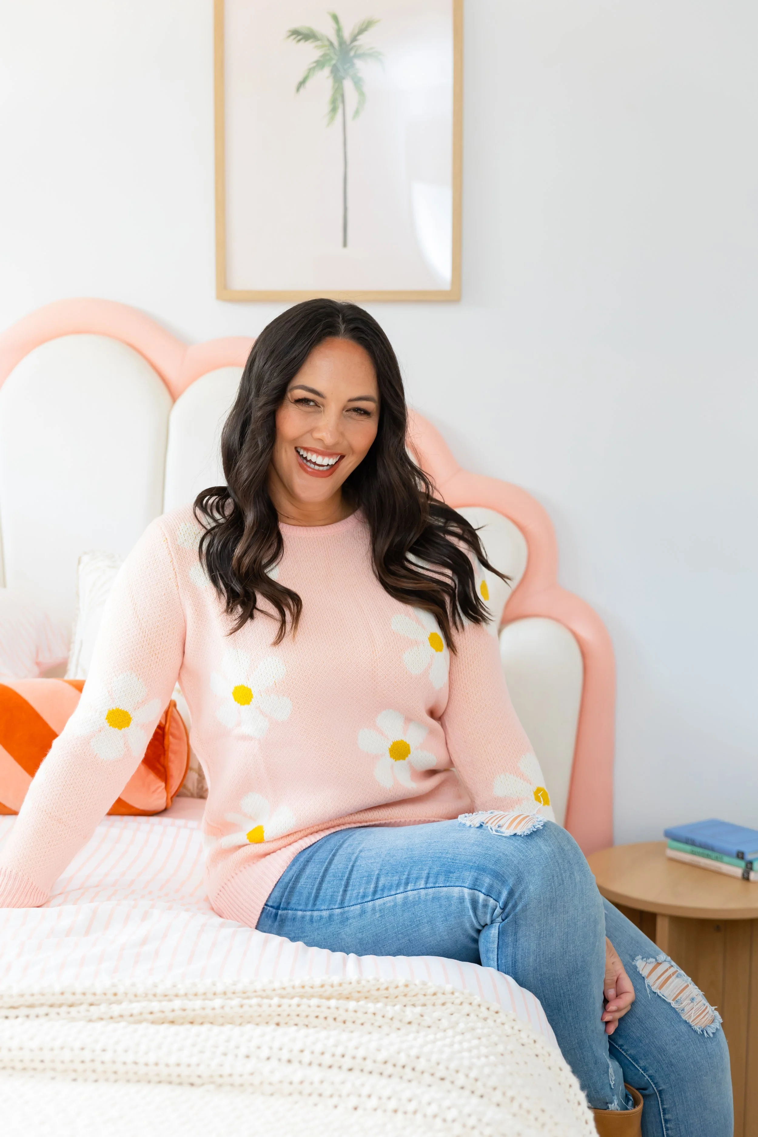 A woman with dark brown wavy hair, wearing a pink sweater with white daisies, sitting on a bed in a brightly decorated room. She is smiling and leaning to her right. The room has a light-colored wall, a framed palm tree picture, and a pink headboard.