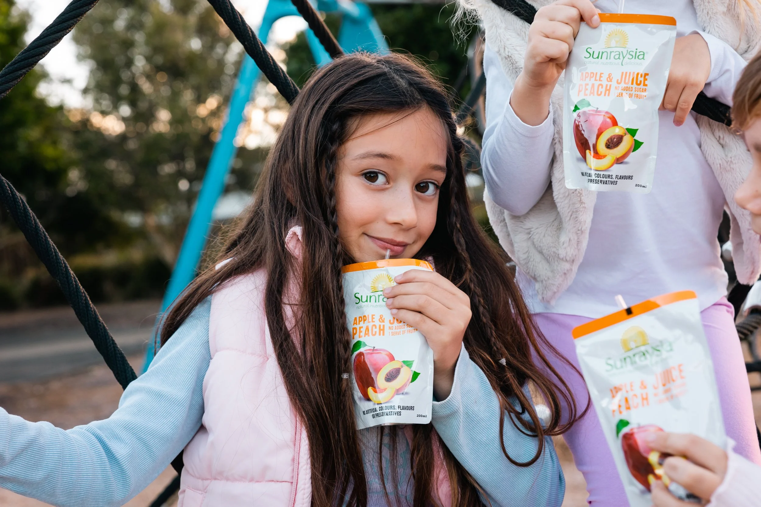 A girl on a playground holding a Sunraysia apple and peach juice pouch, smiling while drinking it, with other children holding similar juice pouches around her.
