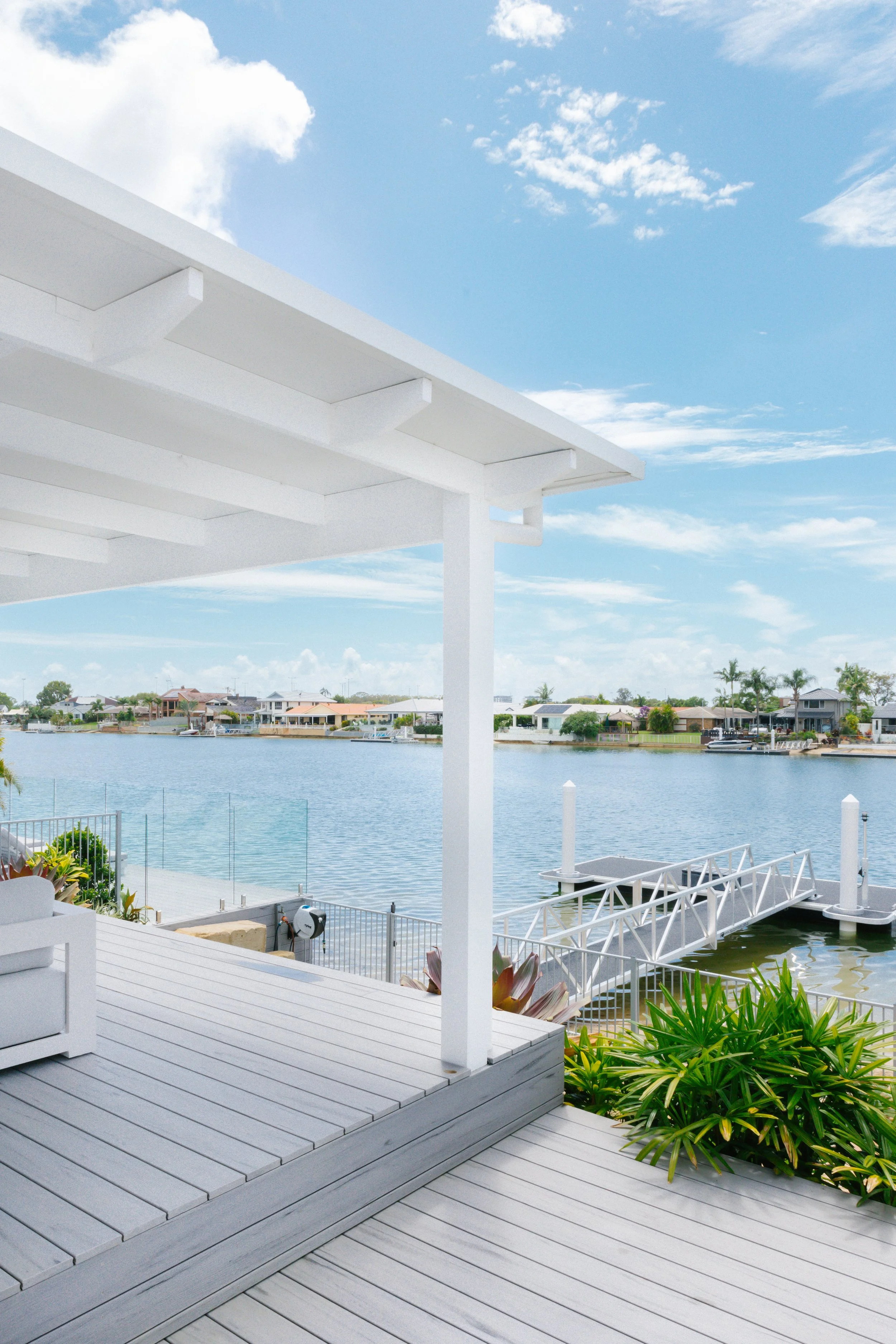 A white modern deck overlooking a calm waterway with houses on the opposite side, a dock with a ramp, and some green plants in the foreground.
