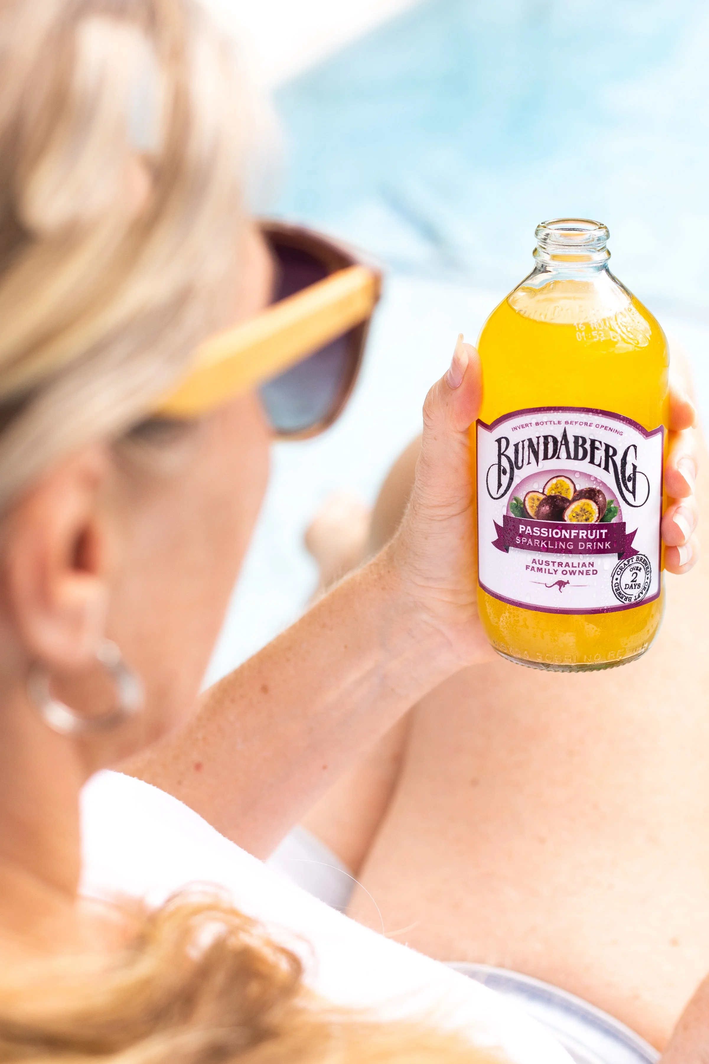 A person with blonde hair and sunglasses holding a bottle of Bundaberg Passionfruit Sparkling Drink.