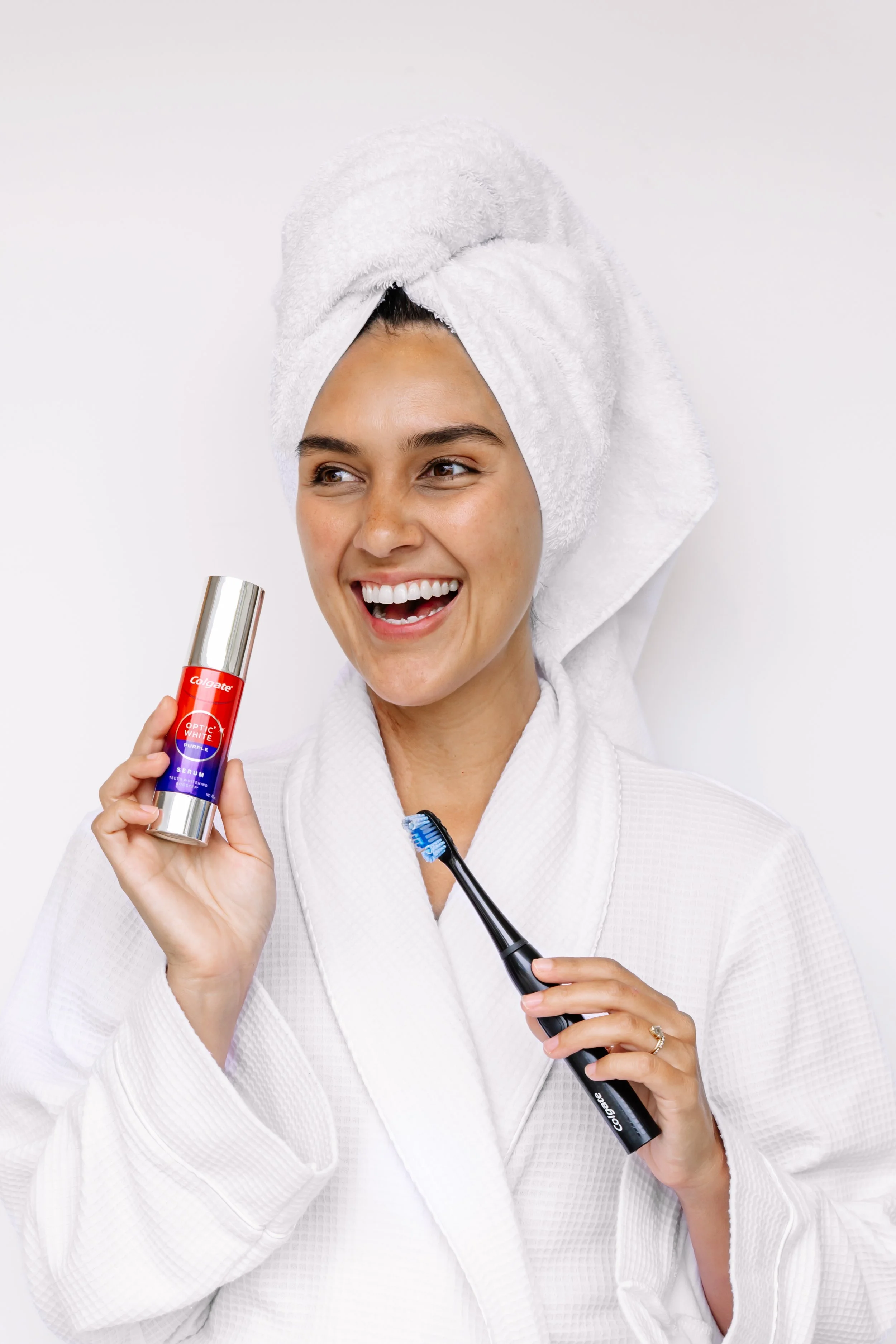 A woman in a white bathrobe with a towel wrapped around her head, holding a toothbrush in one hand and a bottle of whitening serum in the other, smiling.