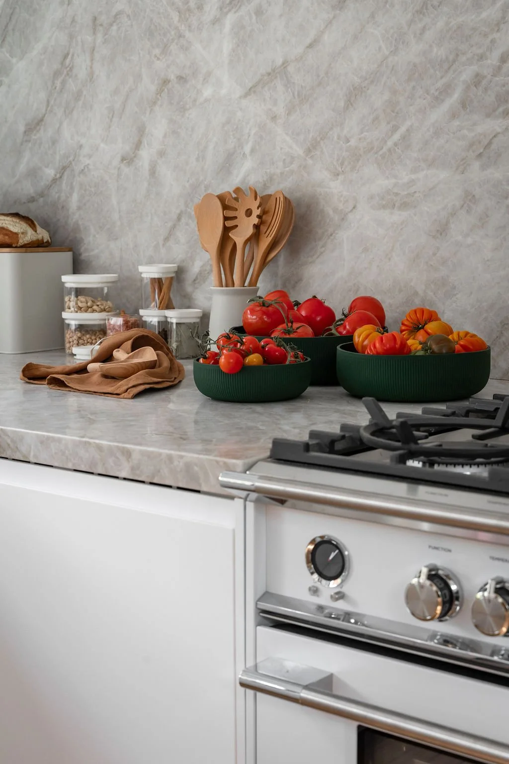 Kitchen countertop with bowls of ripe tomatoes, jars of chickpeas and pretzels, wooden cooking utensils, and a loaf of bread on a marble surface.