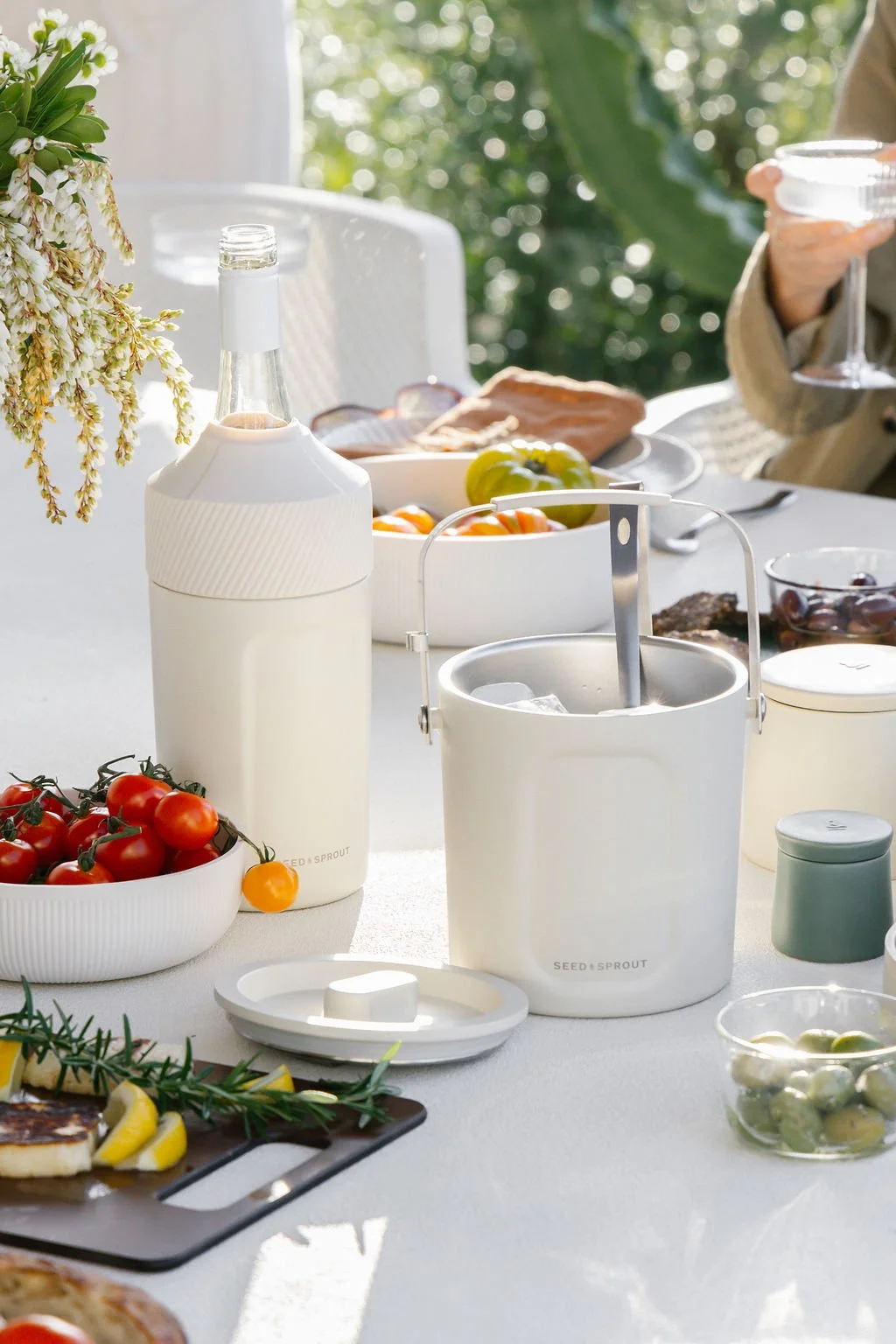 A table set outdoors for a meal with various food items, a bottle, bowls of tomatoes and grapes, and a person holding a glass of wine.