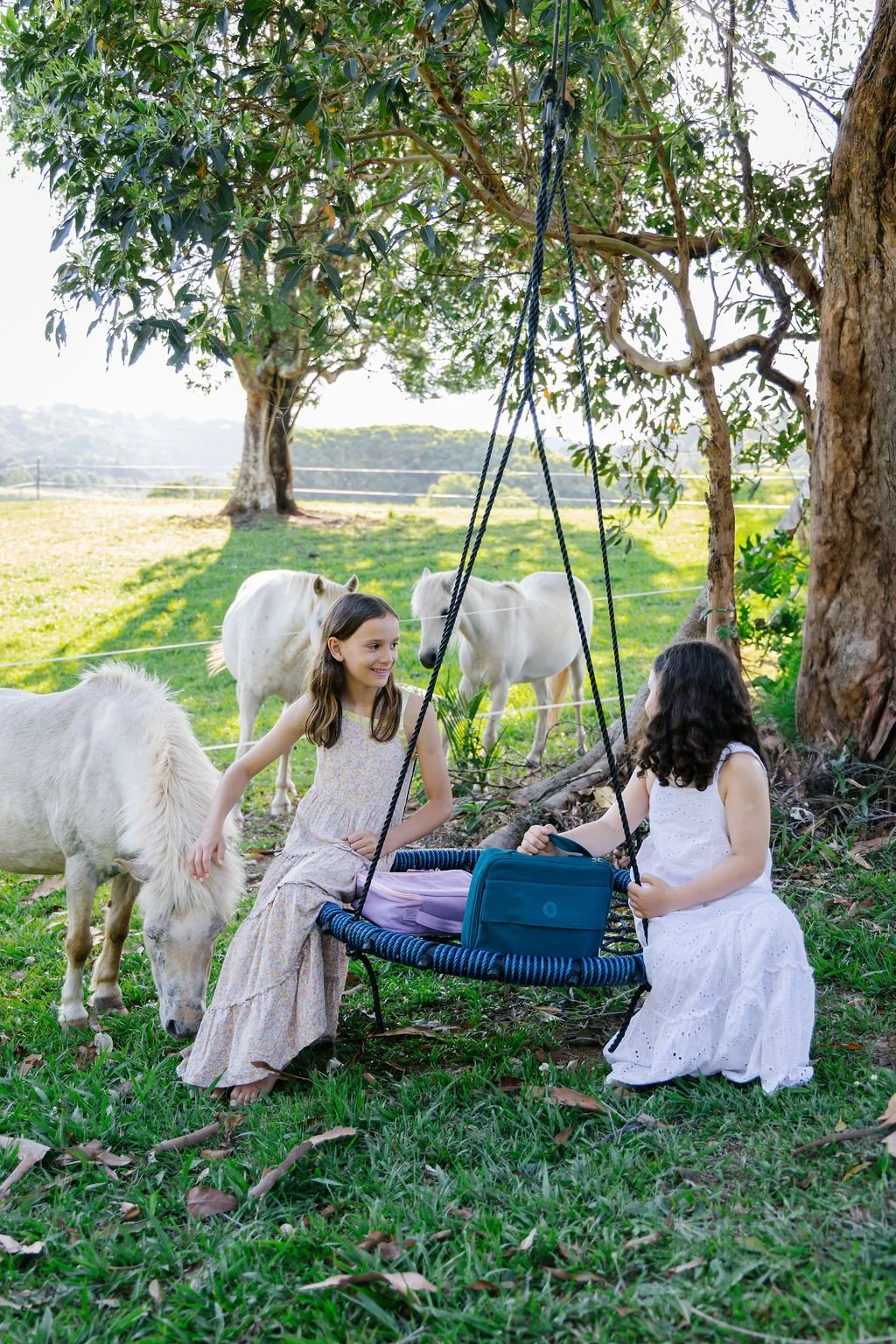 Two young girls in white dresses playing near small white ponies and miniature horses under a large tree in a grassy field.