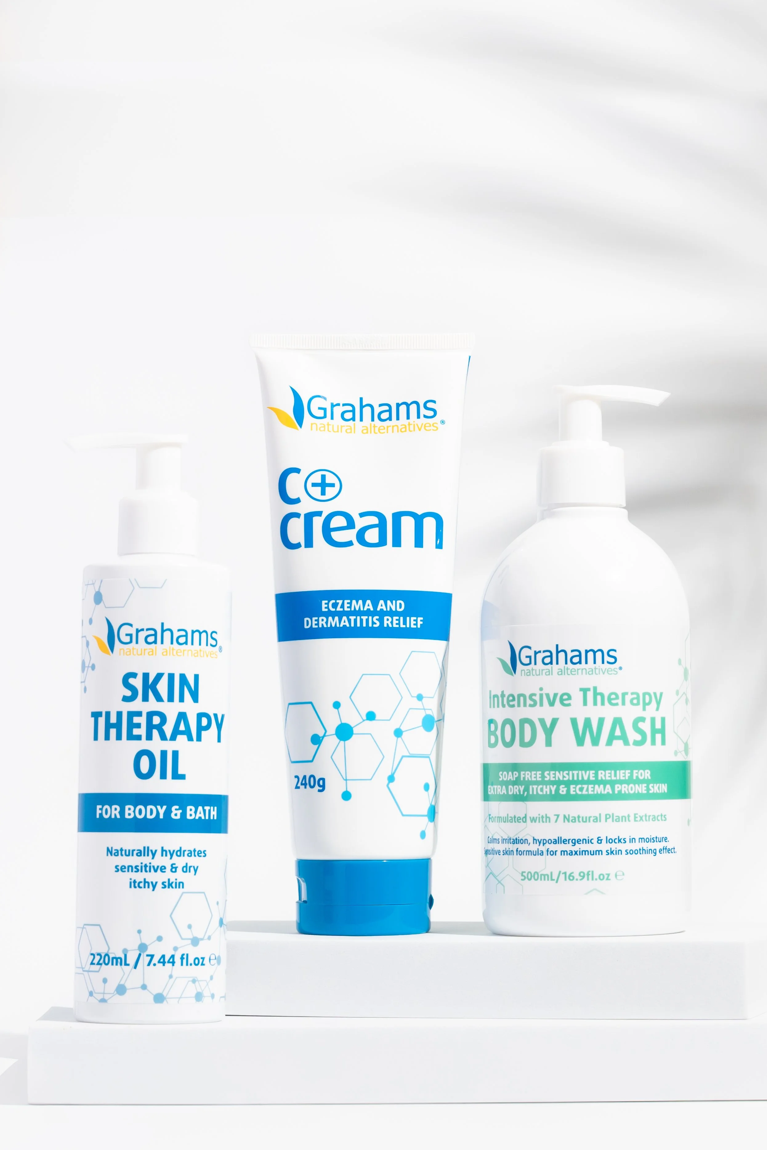 Three skincare products from Grahams with blue and white labels: Skin Therapy Oil, C+ Cream, and Intensive Therapy Body Wash, arranged on a white surface.