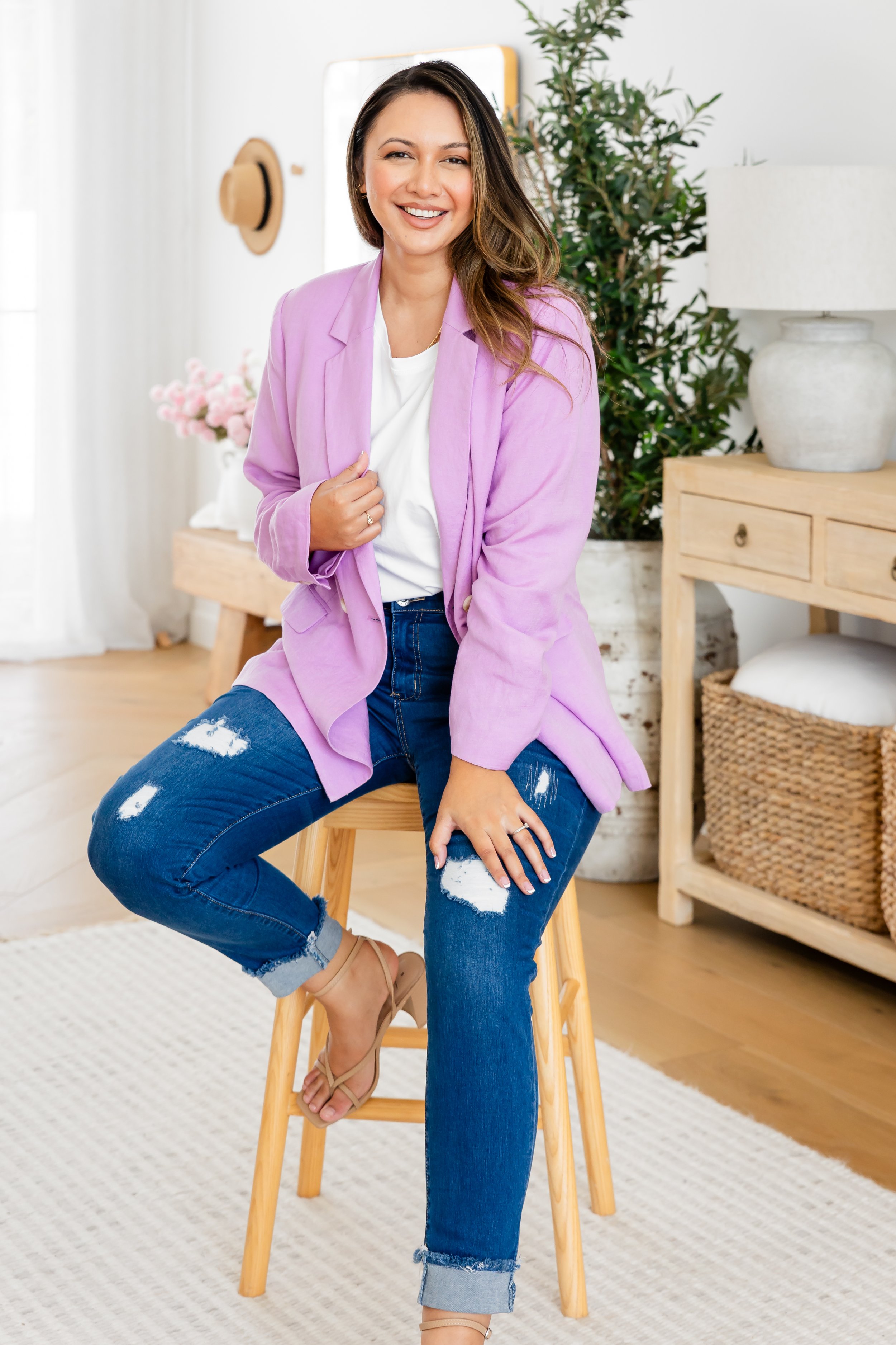 A woman with long brown hair wearing a purple blazer, white t-shirt, and ripped blue jeans sitting on a wooden stool in a bright, cozy room with plants and furniture.