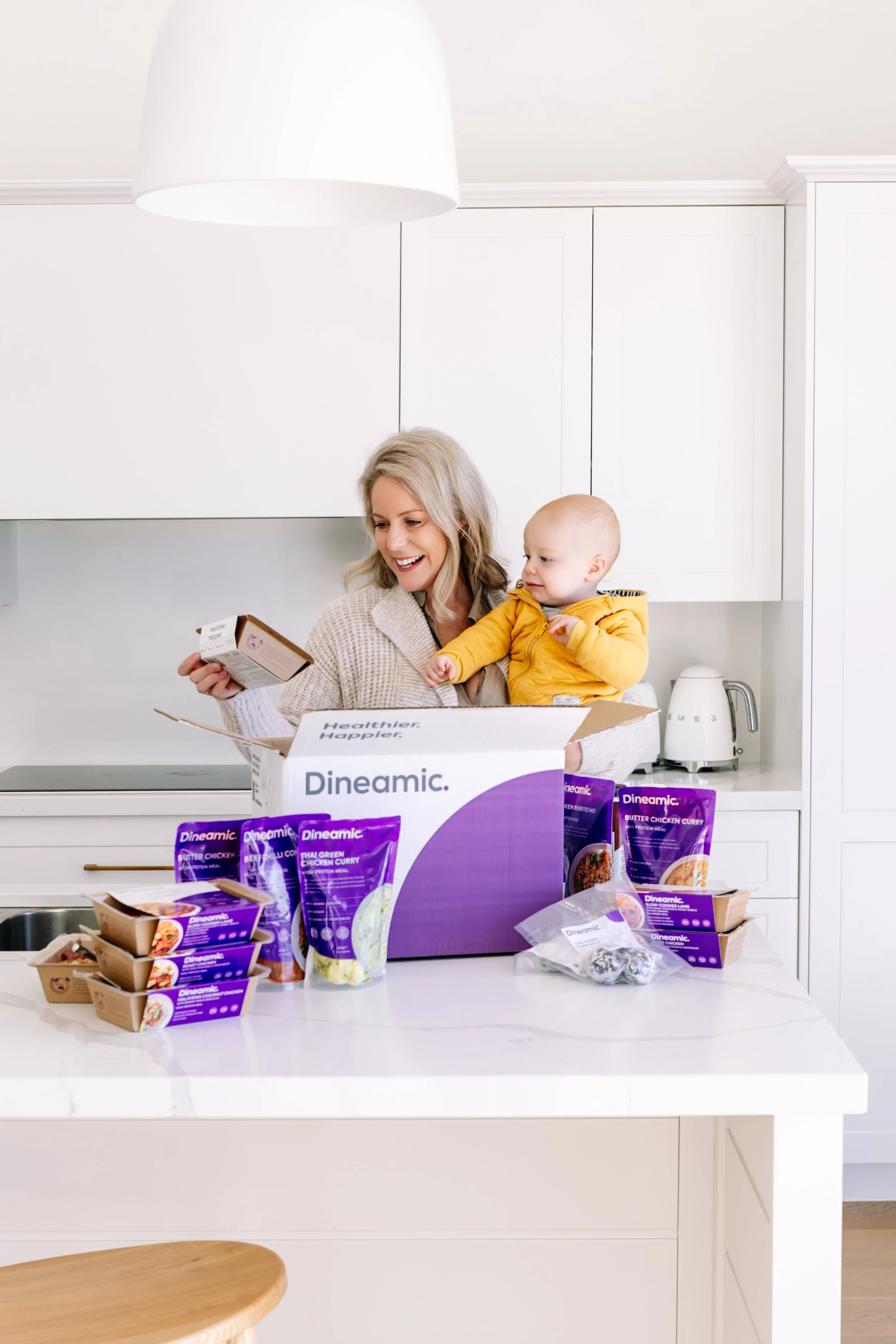 A woman and a young child standing in a kitchen, opening a large box of frozen meals with several packages on the counter.