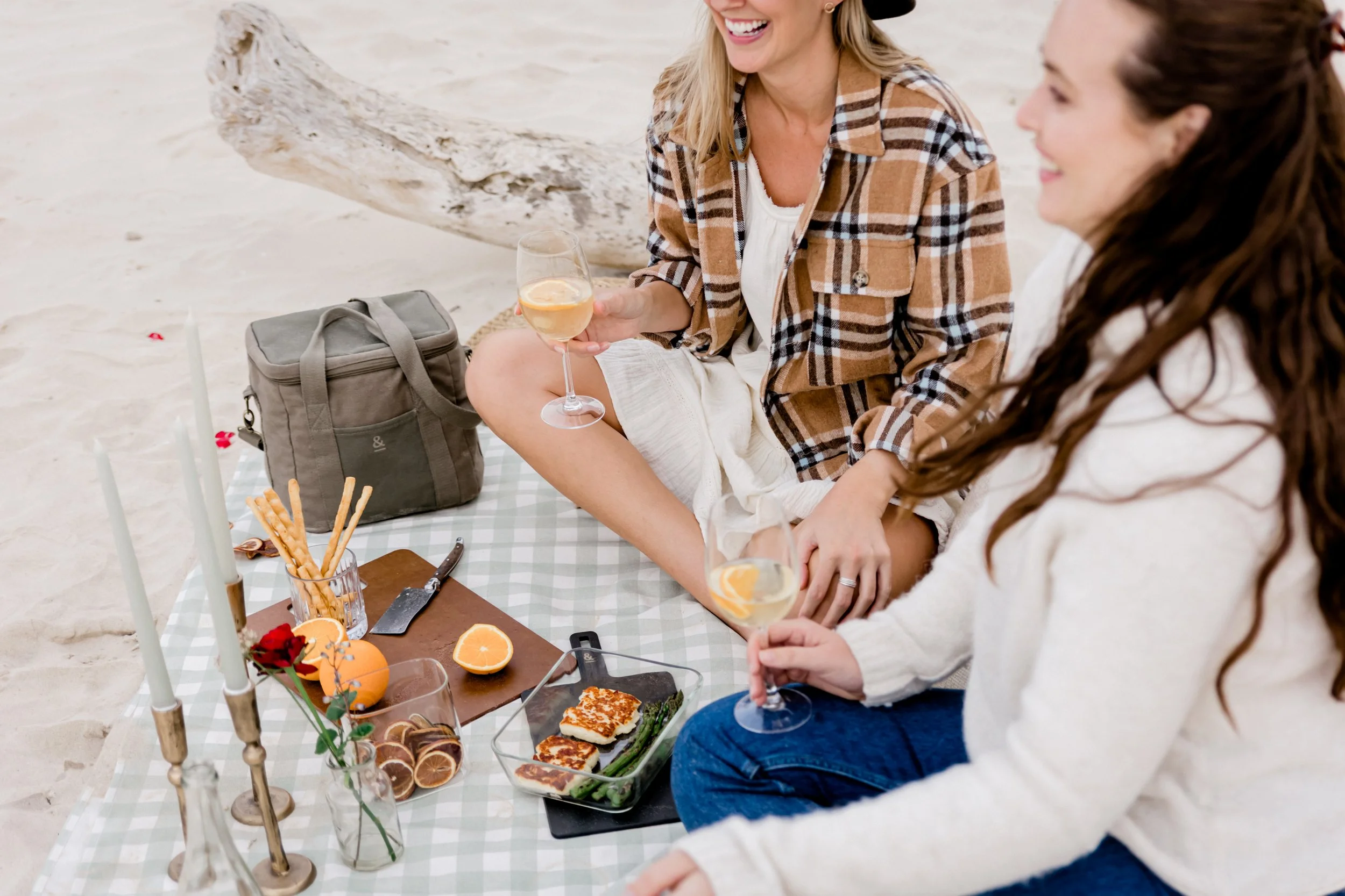 Two women sitting on a checkered picnic blanket on the beach, smiling and holding glasses of white wine with lemon slices, surrounded by snacks, drinks, and candles.
