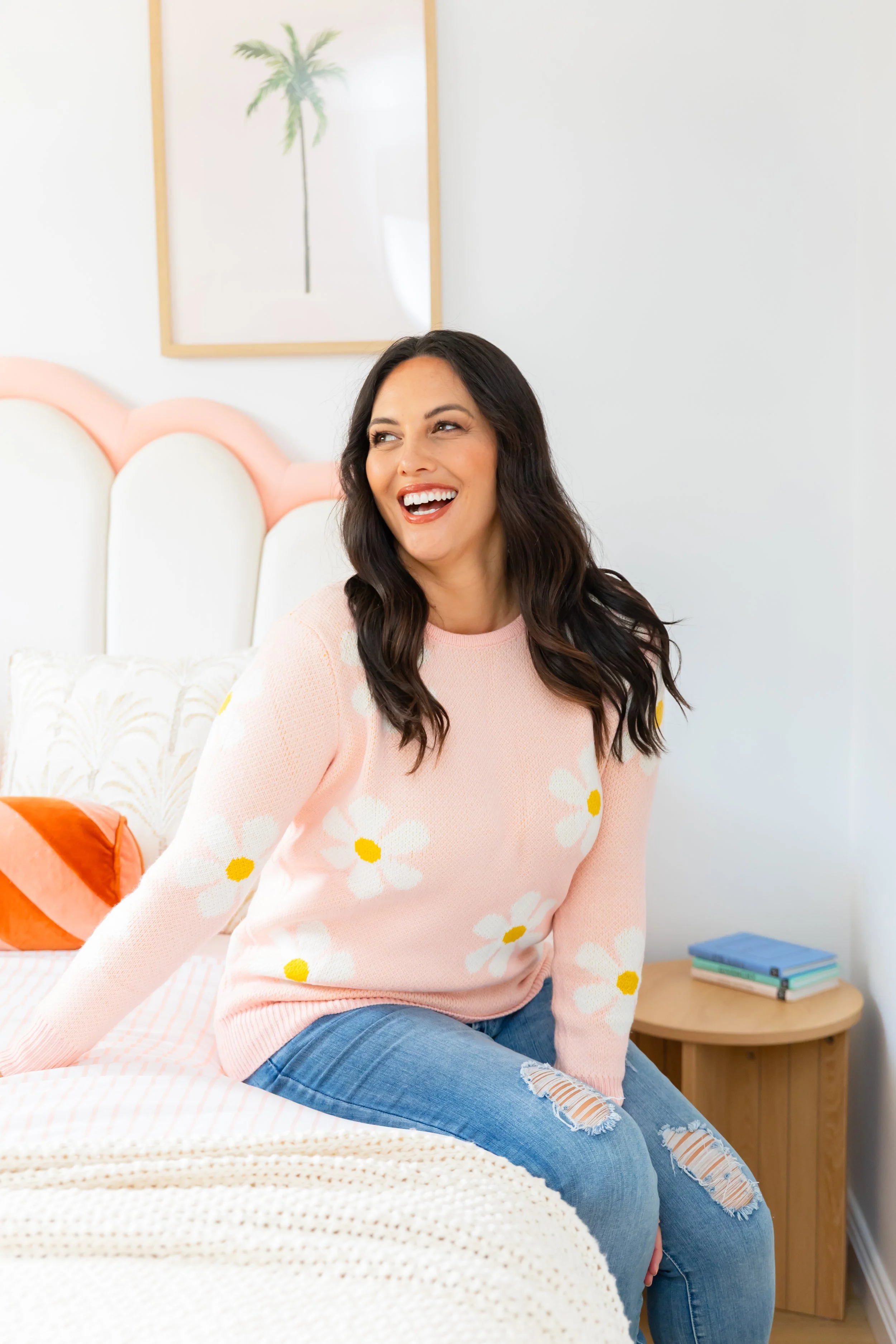 A woman with dark wavy hair sitting on a bed, smiling and looking to the side, wearing a pink sweater with white daisies and ripped jeans, in a bright bedroom with white walls and a framed picture of a palm tree.