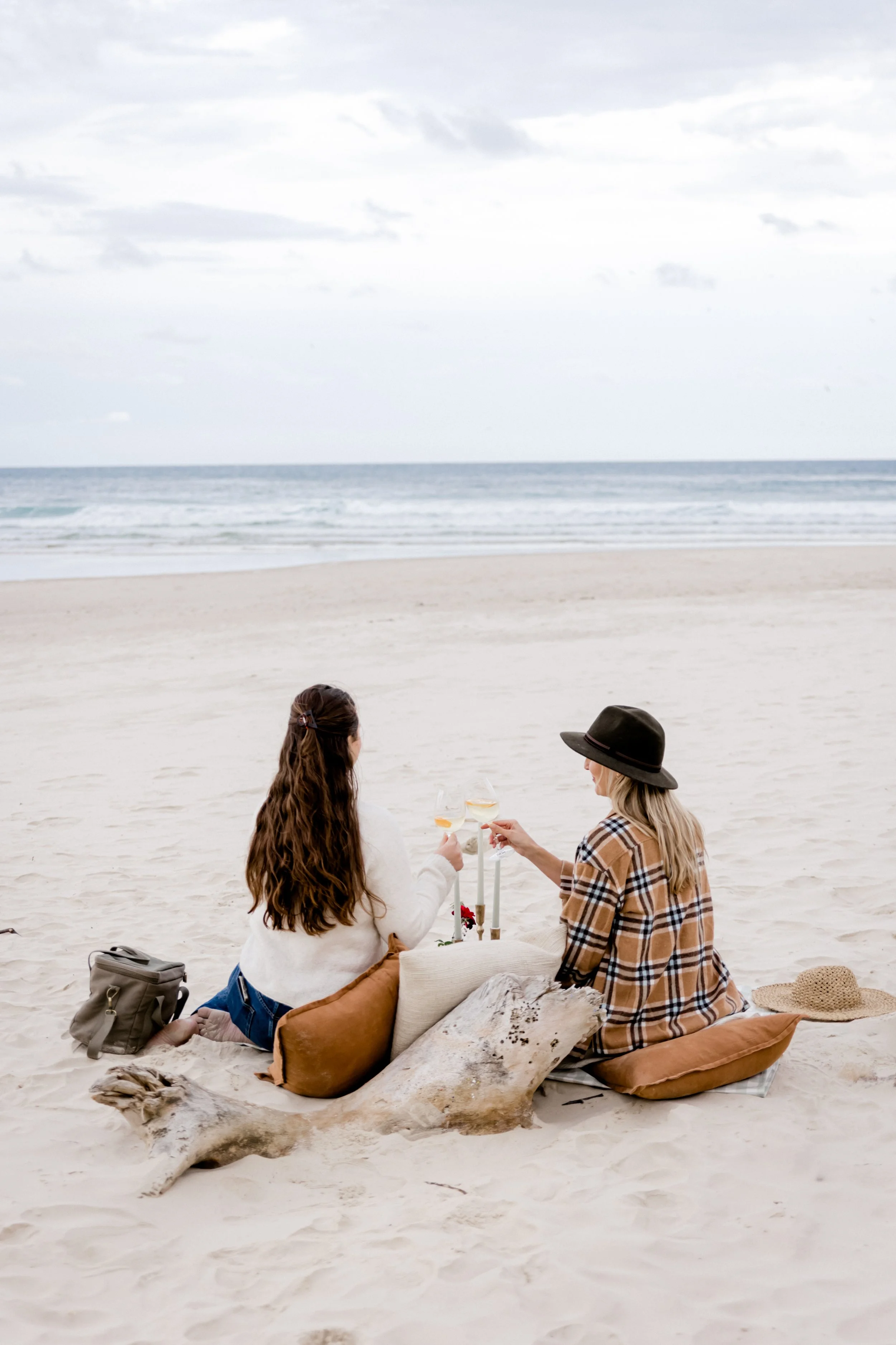 Two women sitting on a sandy beach, facing the ocean, enjoying drinks during an overcast day. One woman has long, wavy brown hair and is wearing a white sweater, while the other is wearing a black hat and a plaid shirt. They have pillows, a straw hat