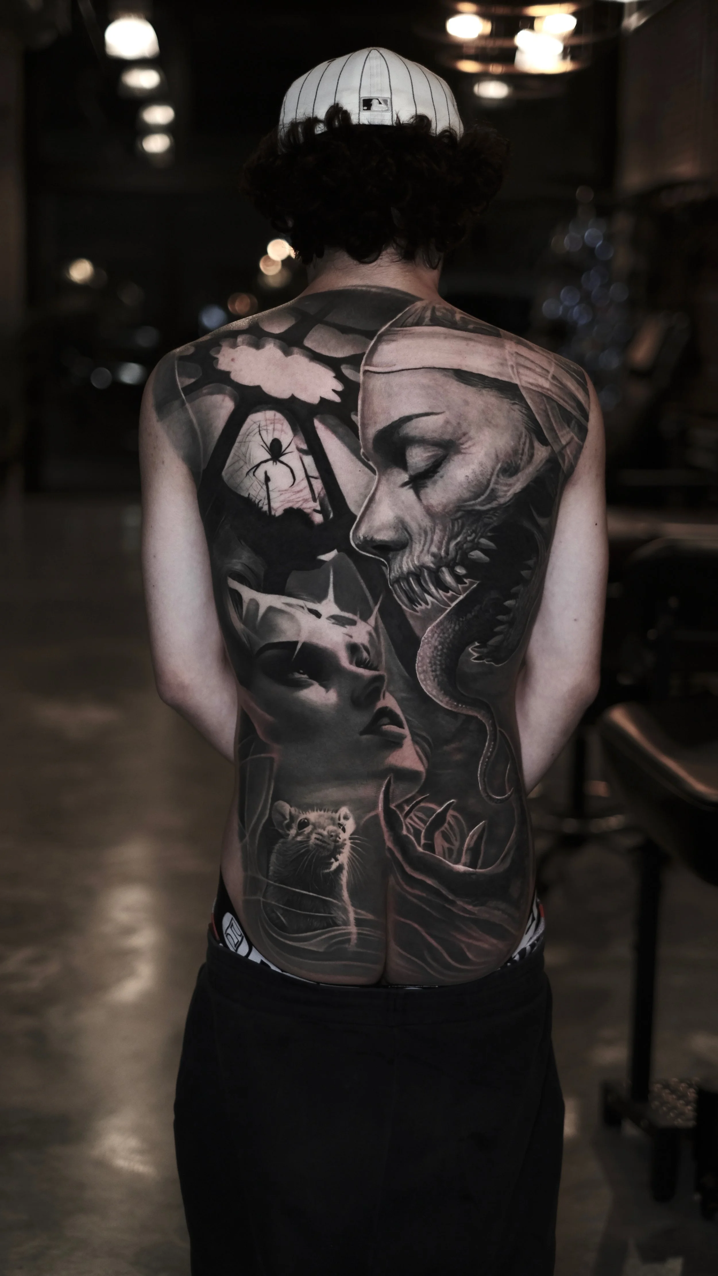 A person with long curly hair, wearing a white baseball cap, shows their back with a large detailed black and gray tattoo covering the entire back. The tattoo features a large skull with a woman's face beneath it, a snake, a girl with a crown, a spid