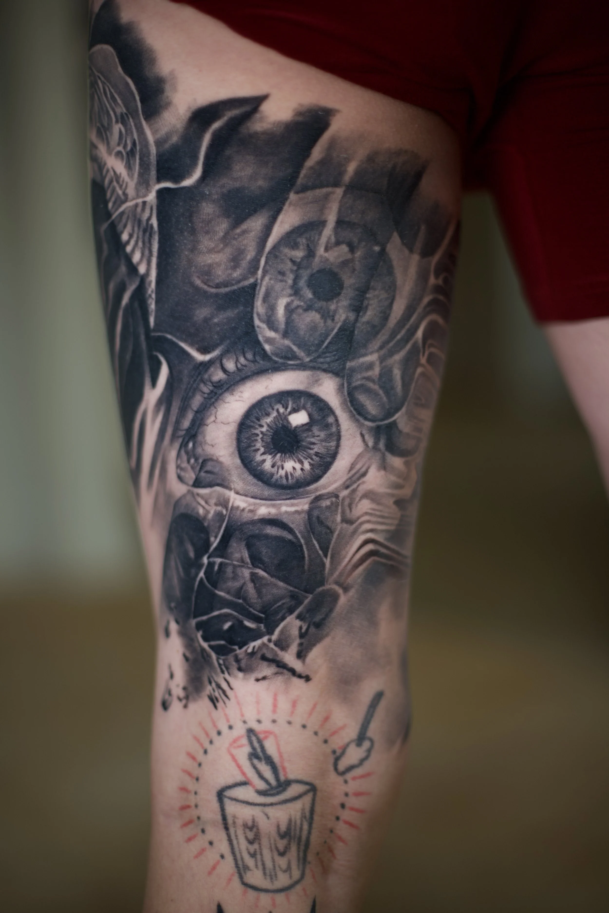 Tattooed arm with a detailed black and gray eye, a skull, and a stylized eye within a circle with rays, alongside other smaller tattoos.