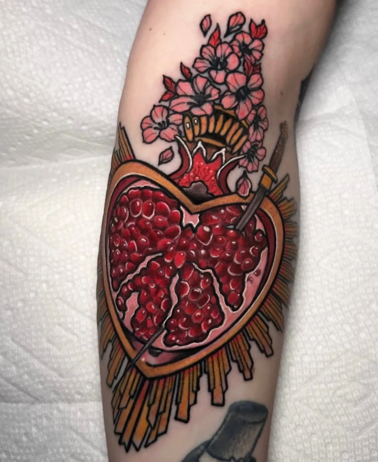 A colorful tattoo of an open pomegranate filled with red seeds, with a bee flying above pink flowers, and a sword piercing through the pomegranate.