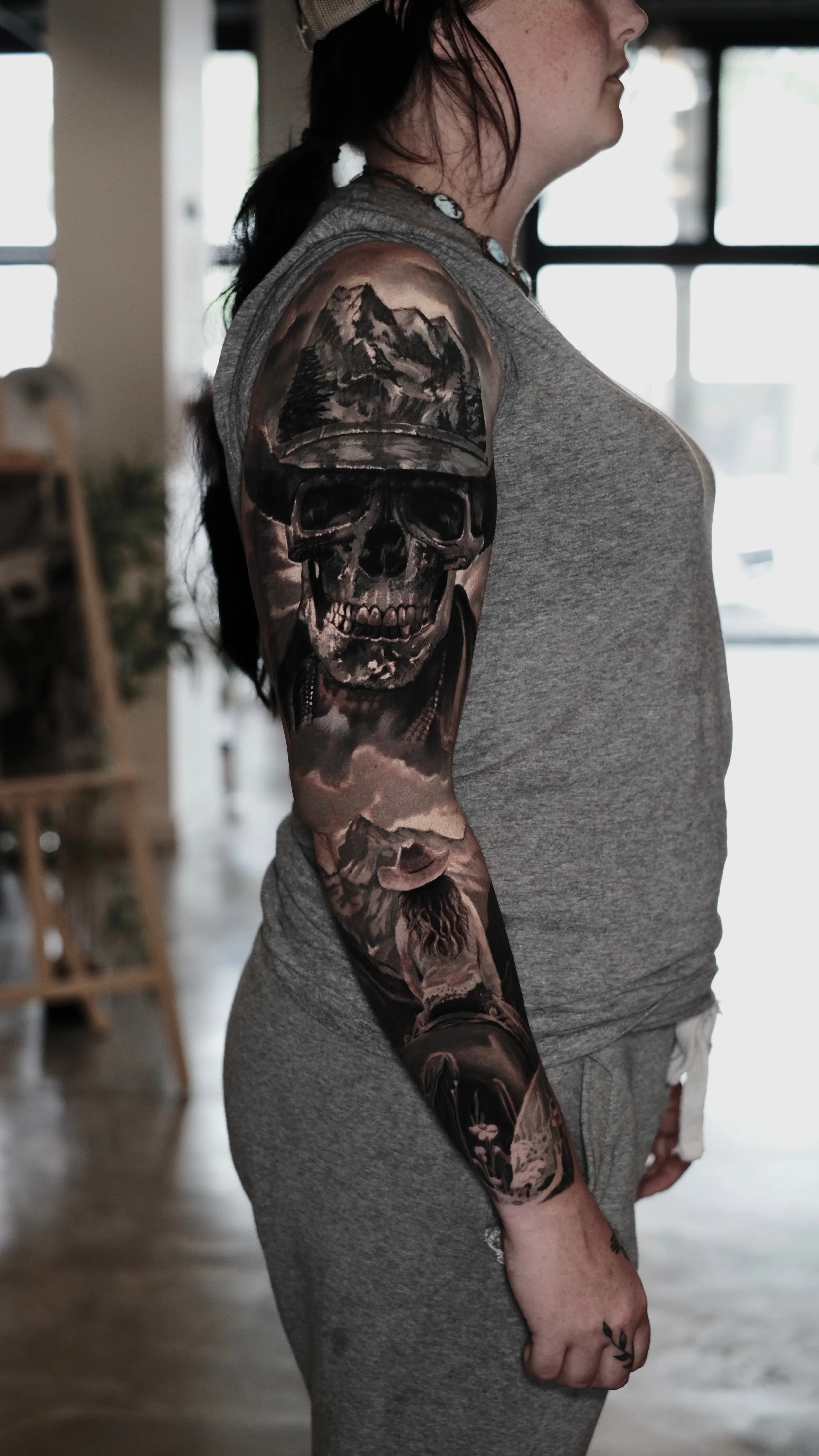 Side view of a person wearing a gray sleeveless shirt, showcasing a detailed black and gray tattoo sleeve featuring mountains, a skull, and a mountain lion. The person has a necklace with shells and has long dark hair.