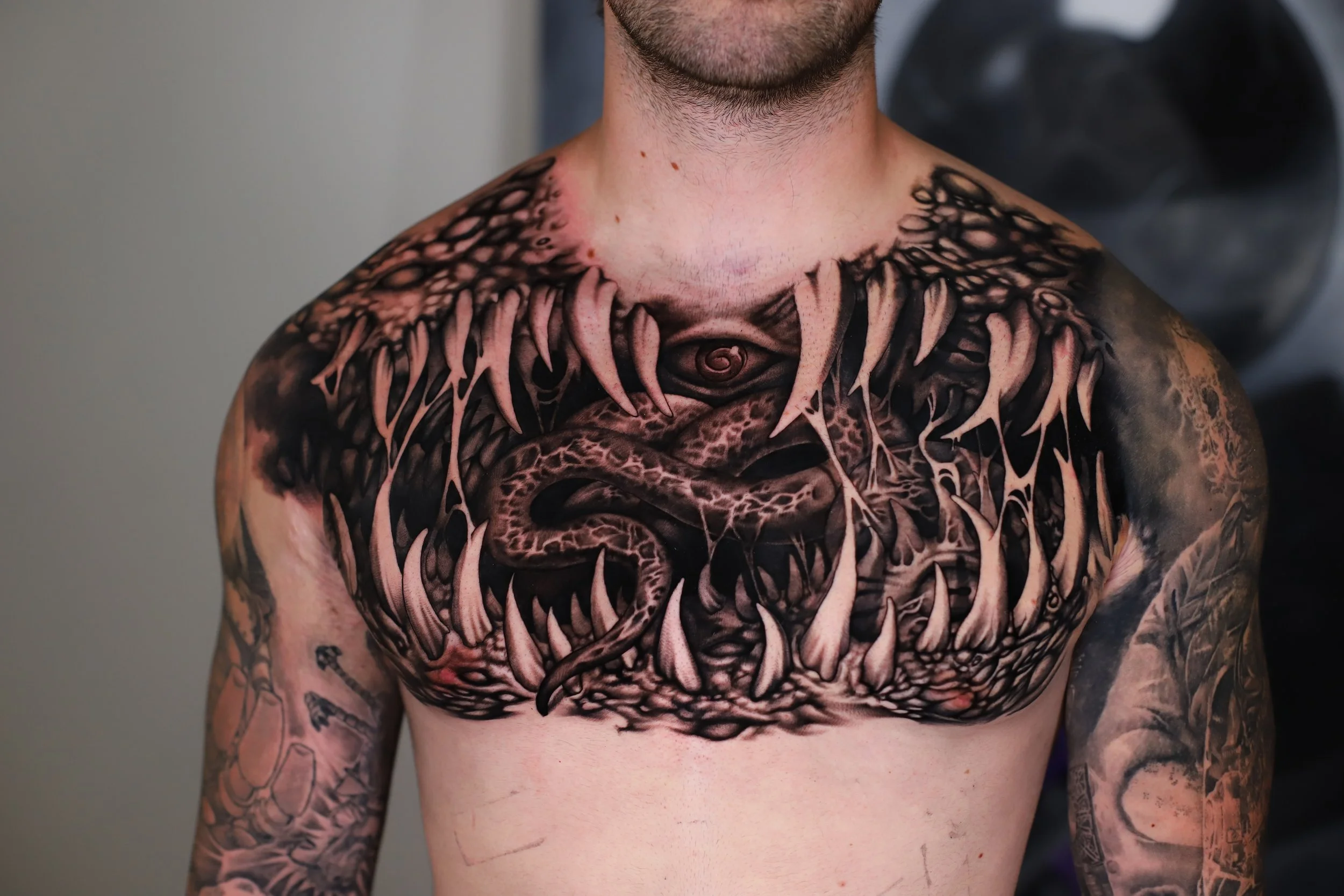 Close-up of a man's upper body with extensive black and pink tattoos, including a large detailed design on his chest containing a central eye and abstract patterns. The man has facial hair and some visible scars on his chest.