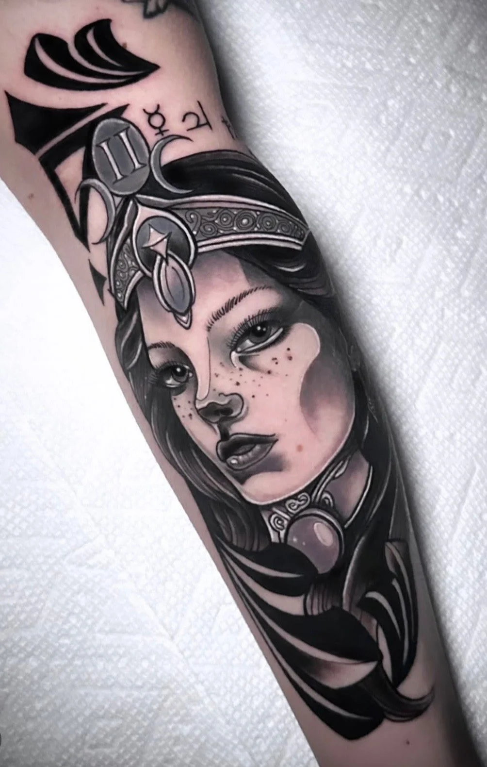 Black and gray tattoo of a woman's face with long hair, featuring freckles, detailed eyes, and a decorative headpiece with geometric patterns.