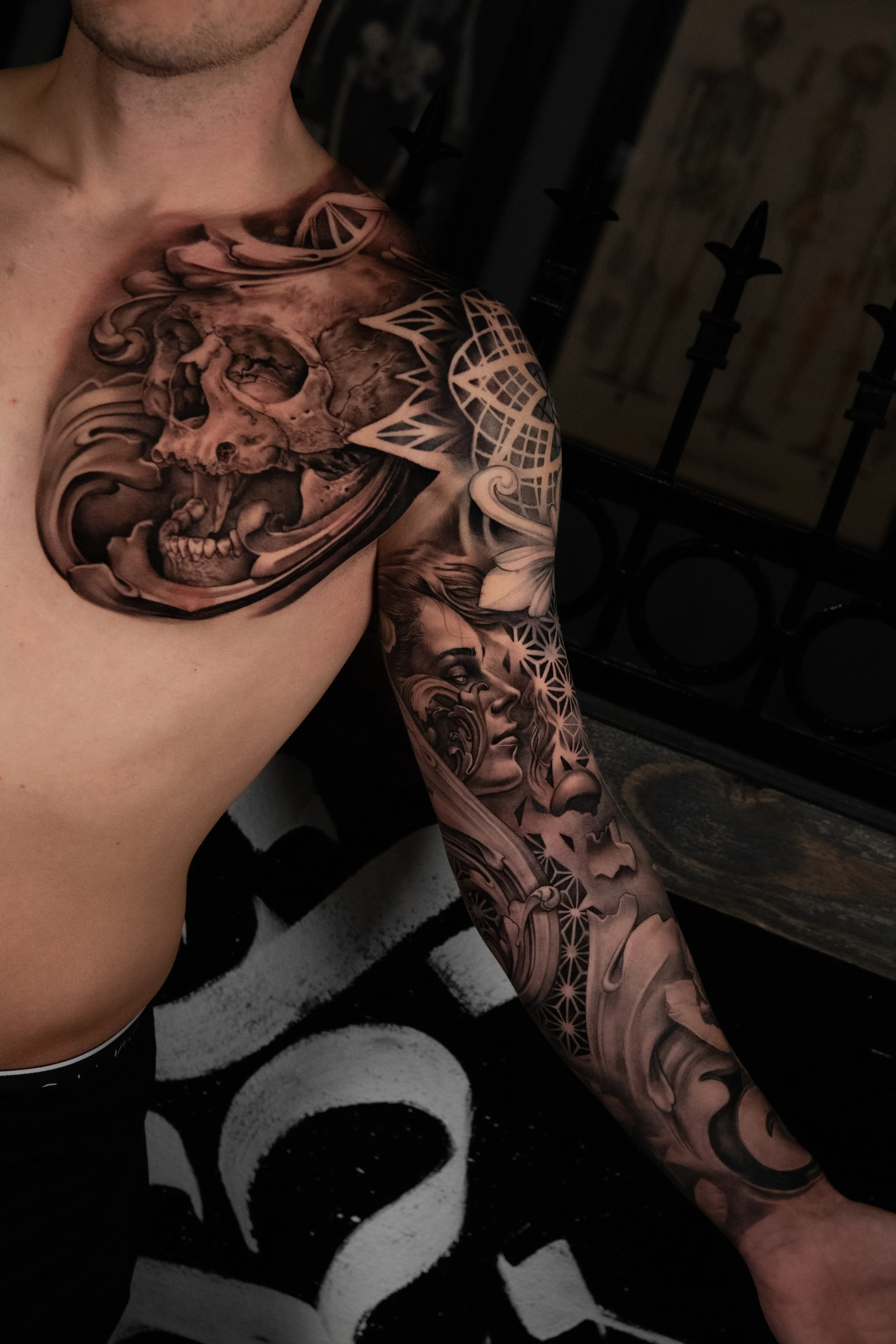 Close-up of a person's upper body with extensive black and gray tattoo sleeves, featuring a realistic skull on the chest and various intricate patterns on the arm.