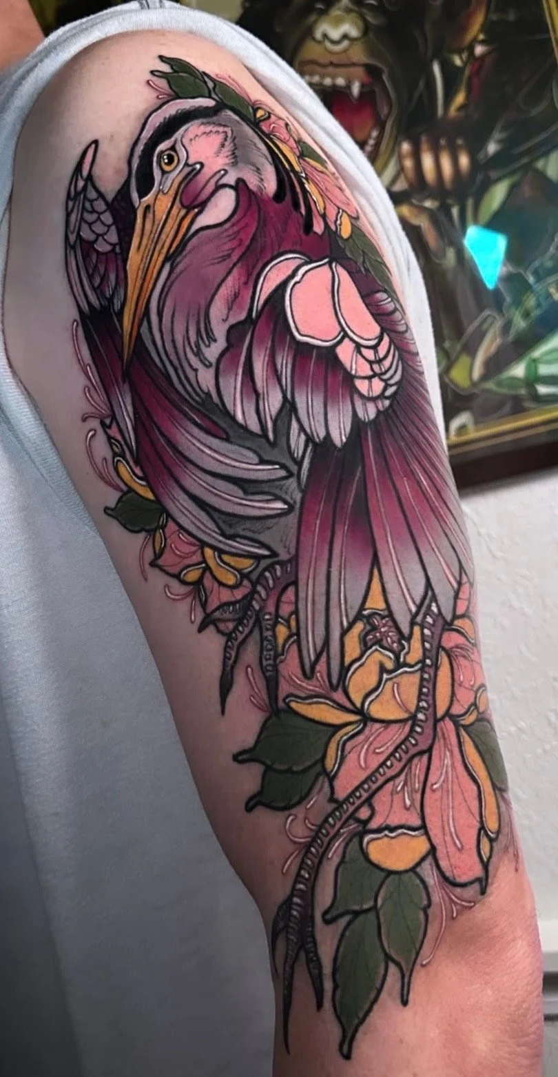 Tattoo of a heron intertwined with pink and orange flowers, with green leaves on an upper arm.