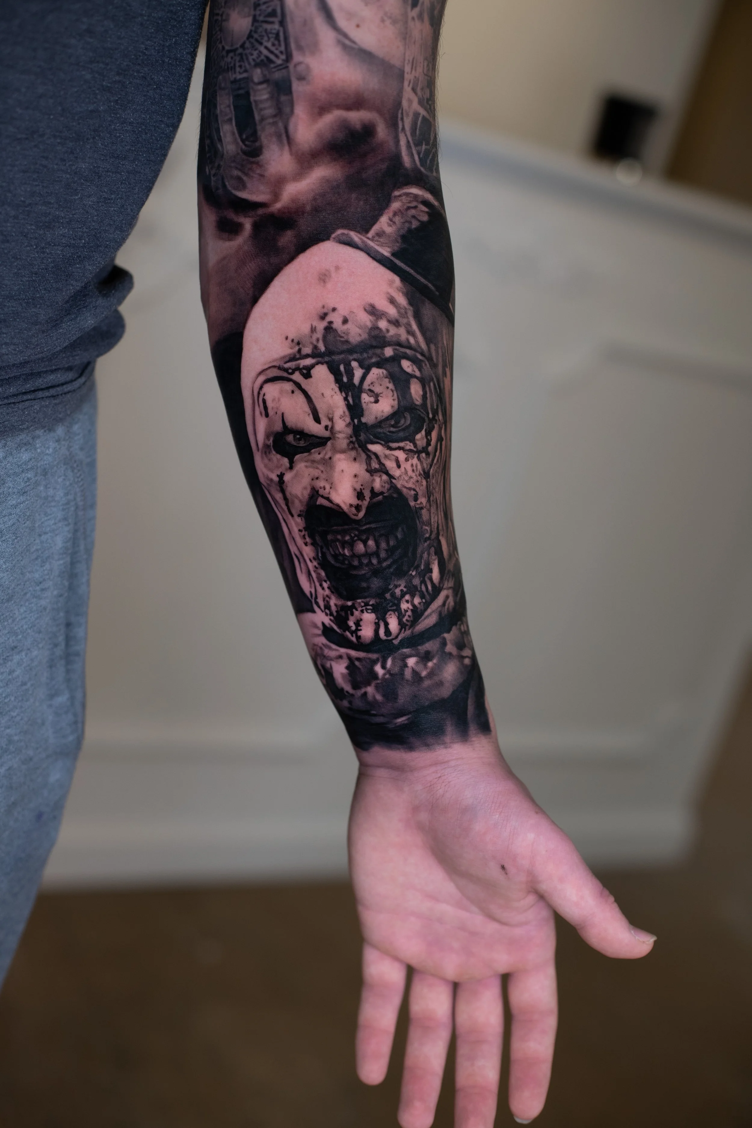 A person with a detailed black and gray tattoo sleeve on their left arm. The tattoo features a skull, a face, and other intricate designs. The person has light skin and is holding their hand in a relaxed position with fingers slightly spread. The bac