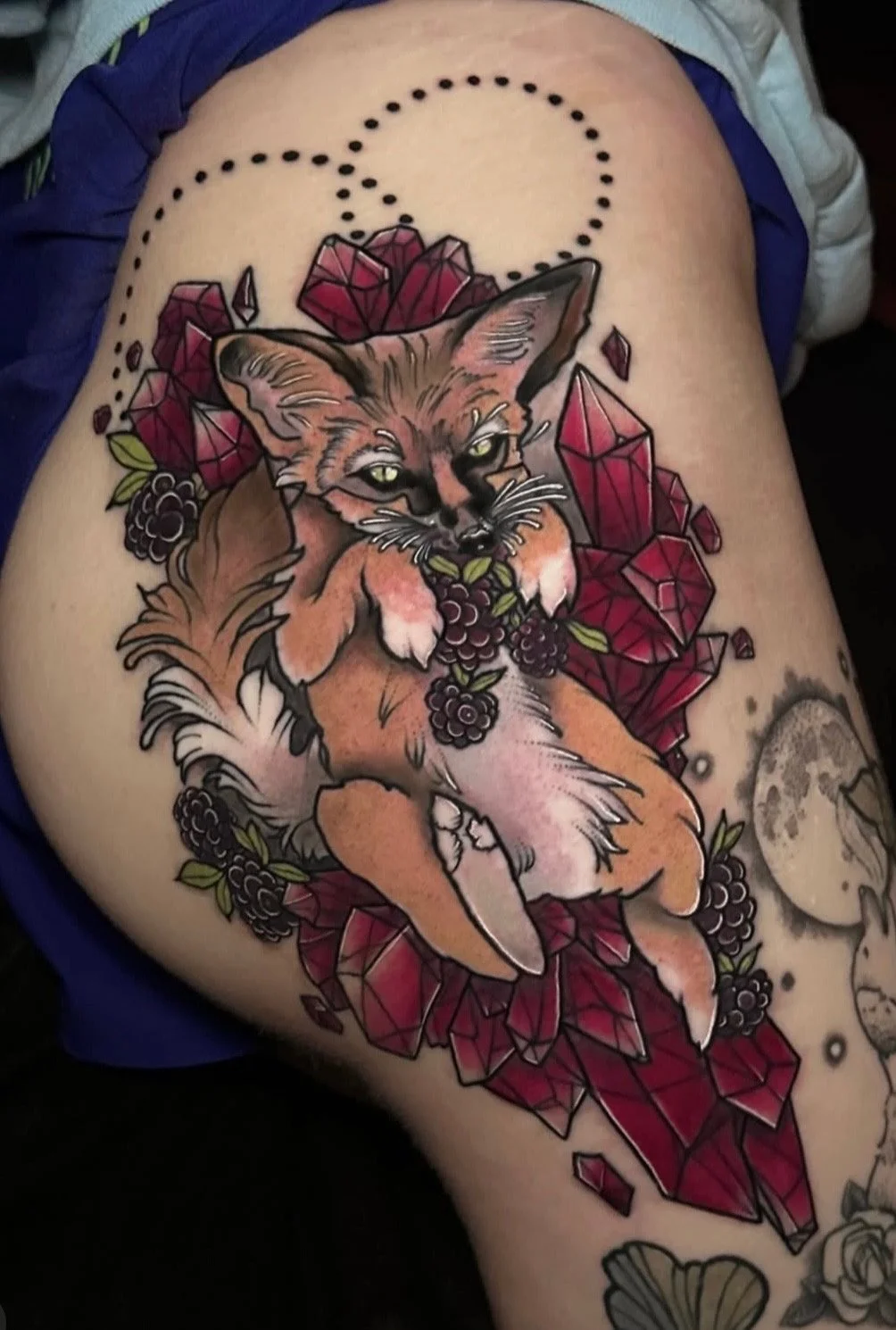 Tattoo of a fox surrounded by red gemstones, violets, and leaves on a person's thigh.