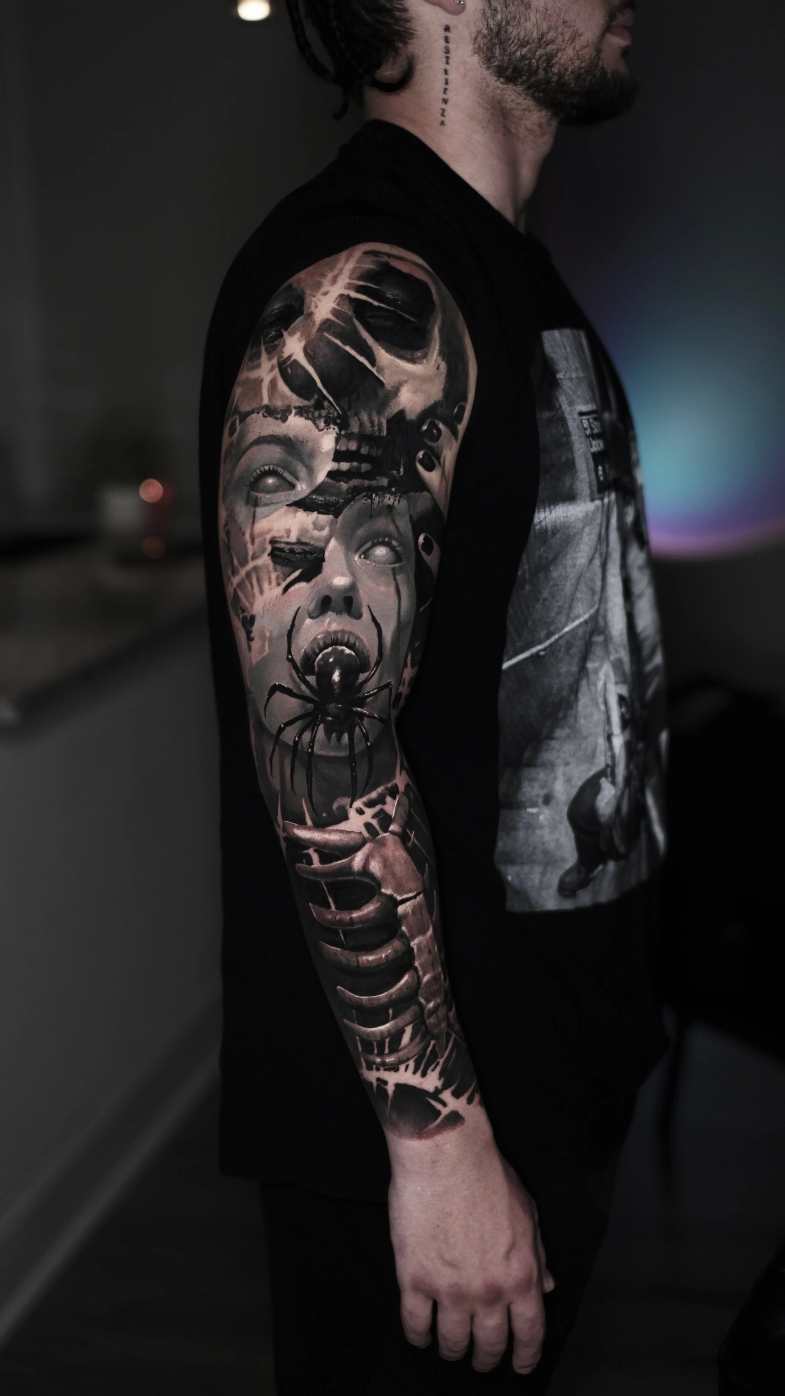 A person with black hair, a choker, and a tattoo on their neck. They are wearing a black sleeveless shirt, displaying a detailed black and gray tattoo sleeve featuring skulls, faces, and an insect on their arm.