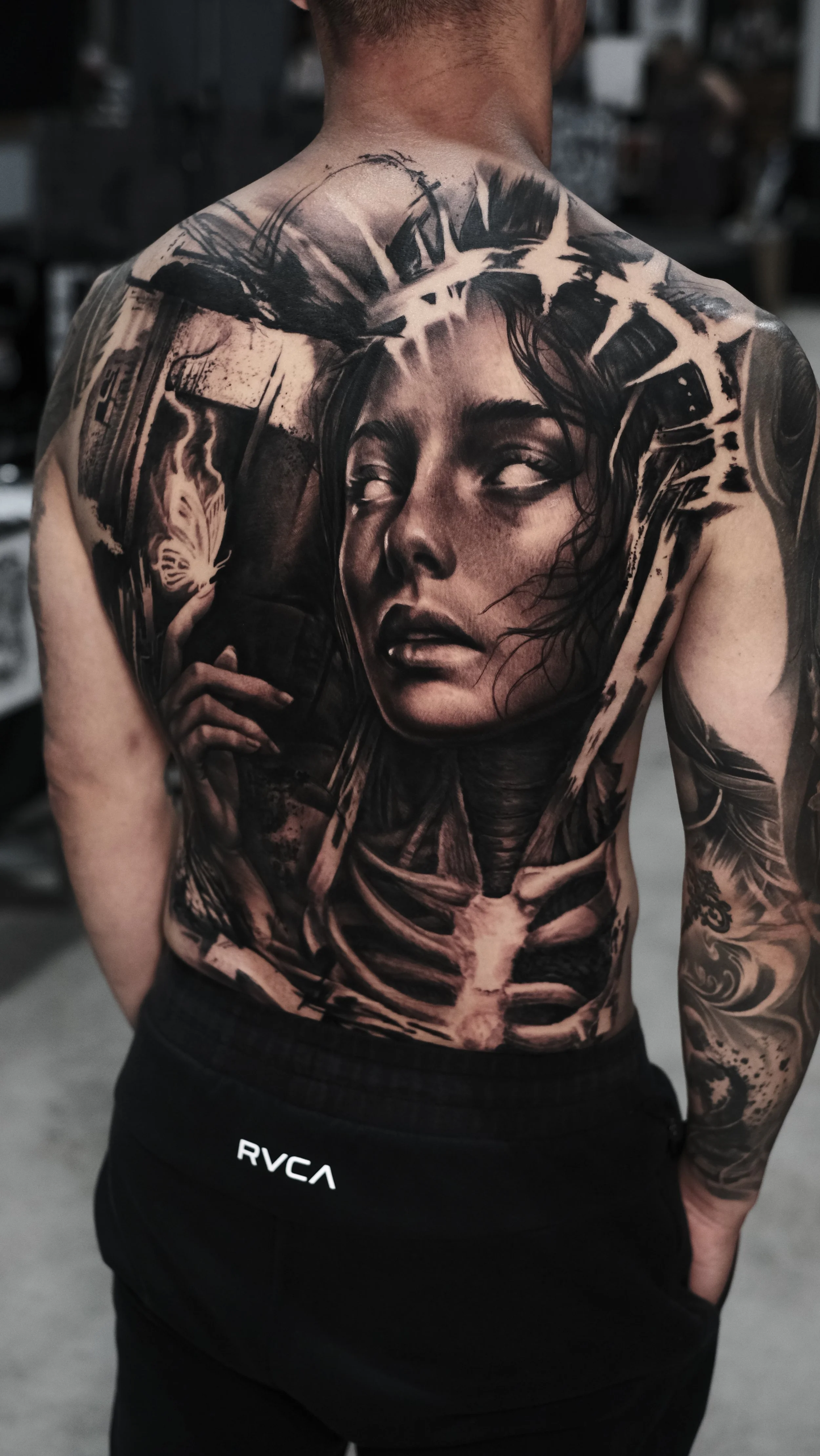 A person with an upper body tattoo featuring a realistic portrait of a woman's face, with dark hair, looking slightly upwards, surrounded by abstract and mechanical design elements, in black and gray ink.