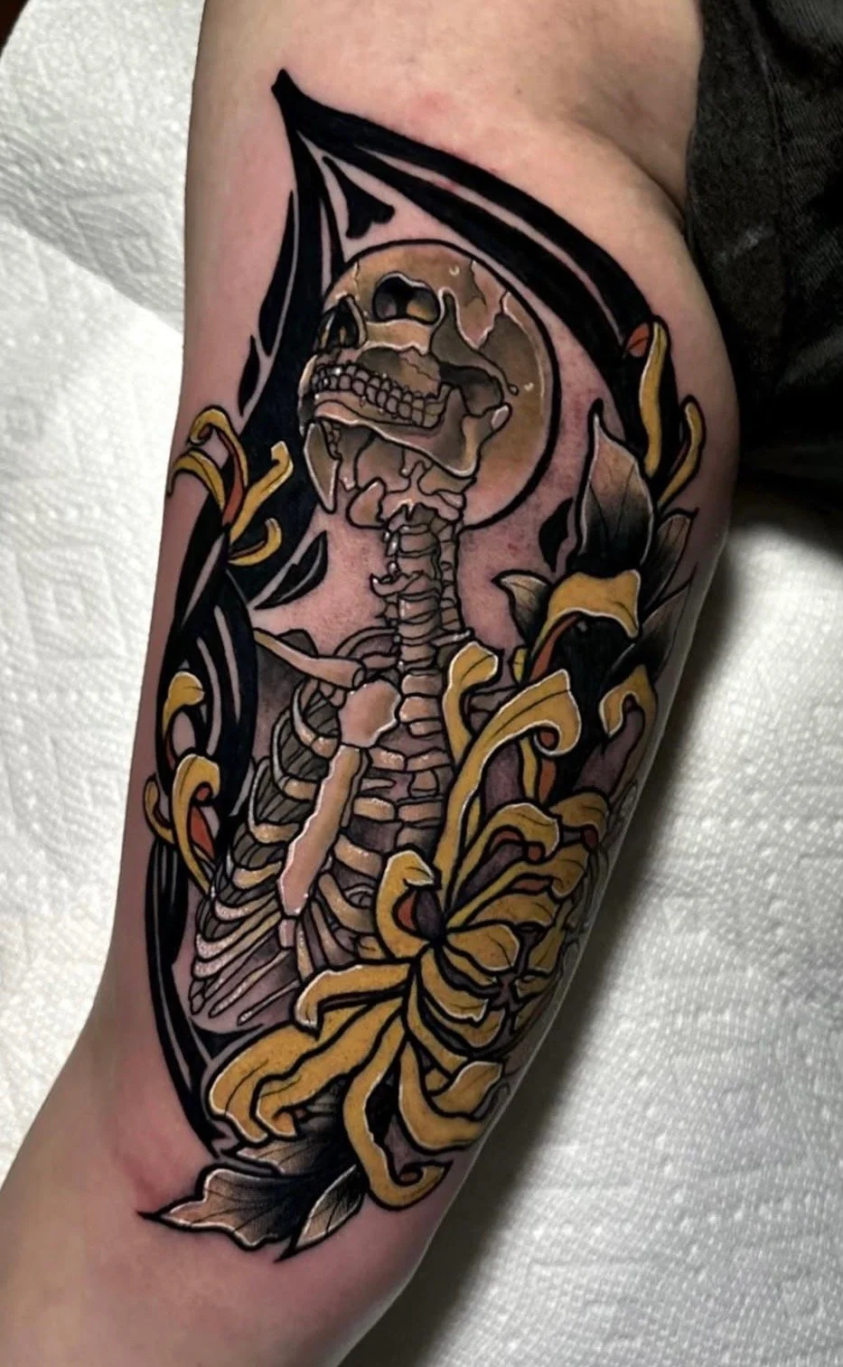 Tattoo of a human skeleton with a skull, surrounded by a black and yellow floral design.