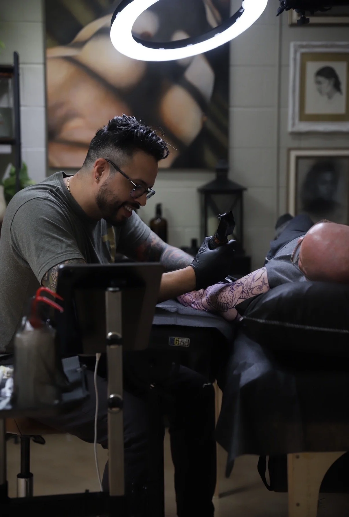 A tattoo artist is working on a client's arm in a tattoo studio, with a large portrait on the wall behind them.
