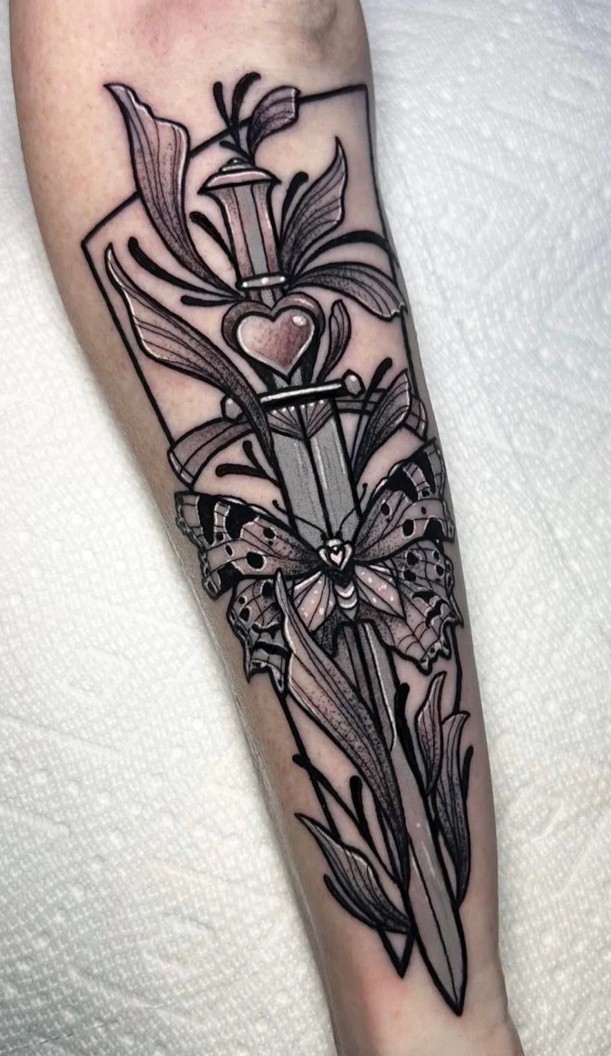 Tattoo of a large sword with leaves, a butterfly, and a heart, designed with black outlines, shading, and some pink details.
