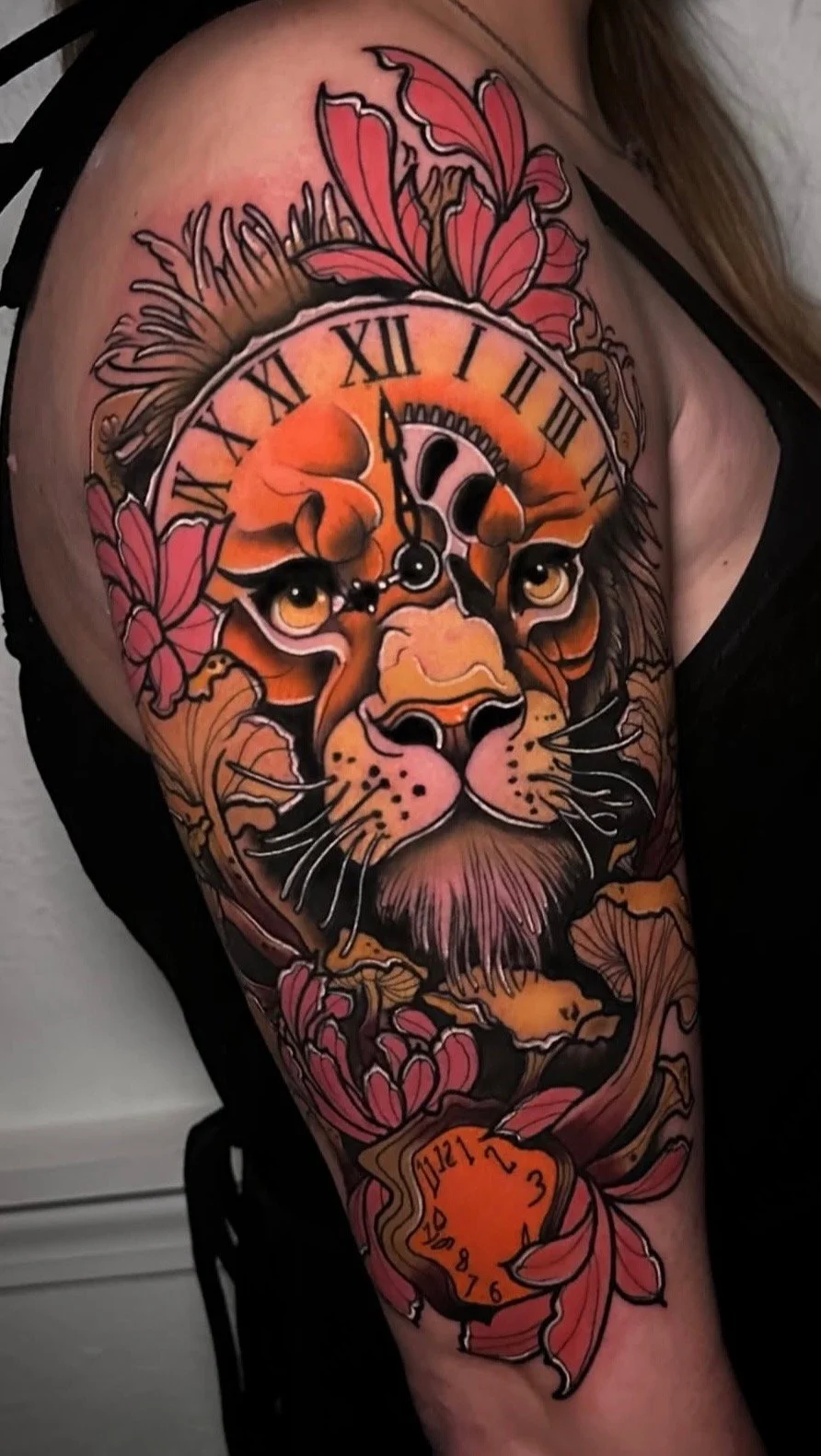 Colorful tattoo of a lion's face with a clock on its forehead, surrounded by pink flowers and leaves, with a small orange clock at the bottom.